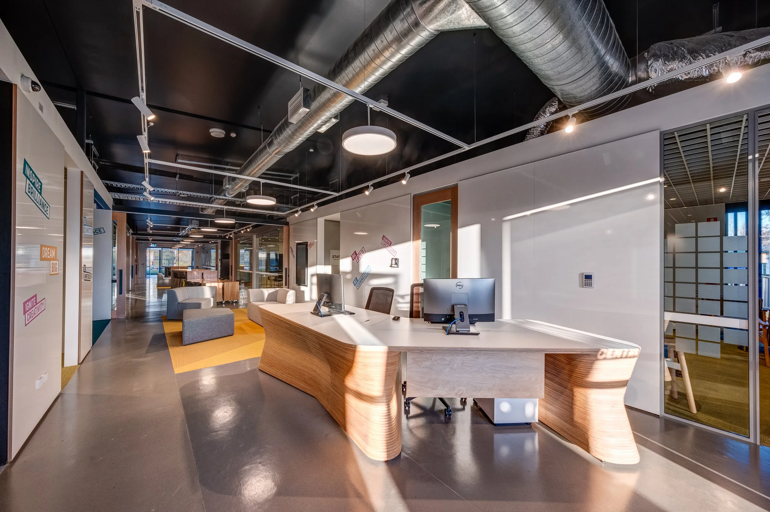 Modern office reception area with a curved wooden desk, computer monitors, and seating area with sofas, illuminated by ceiling lights and featuring colorful motivational posters on the walls.