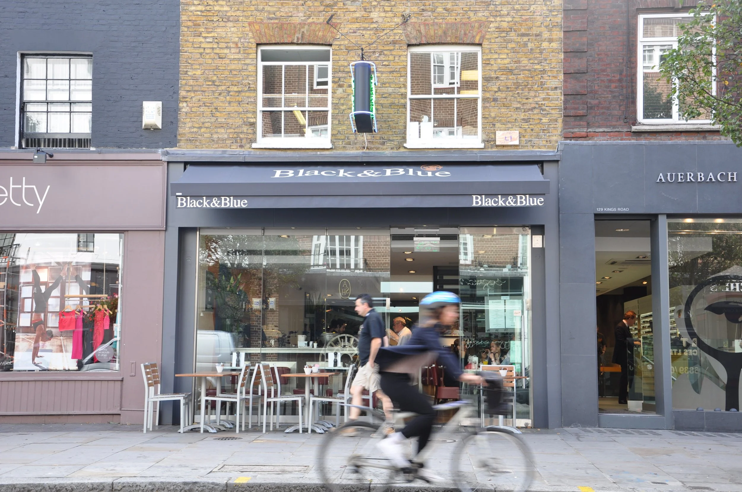 Street view of a commercial building with storefronts, people walking, and a cyclist passing by. The storefronts include Black & Blue and Auerbach, with outdoor seating visible in front of Black & Blue.