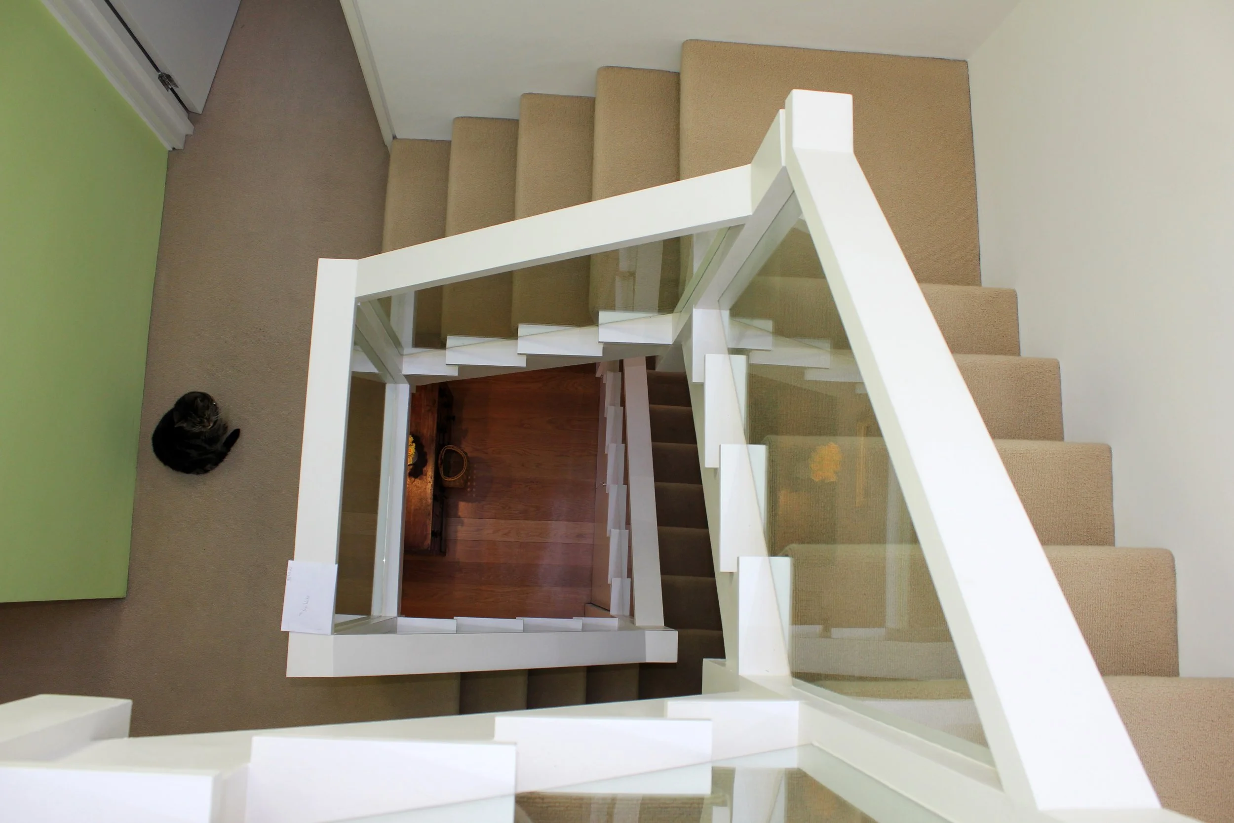 A staircase with beige carpeted steps and white railing view from above, with a green wall on the left and a white wall on the right. A cat is lying on the beige carpet near the bottom left corner.