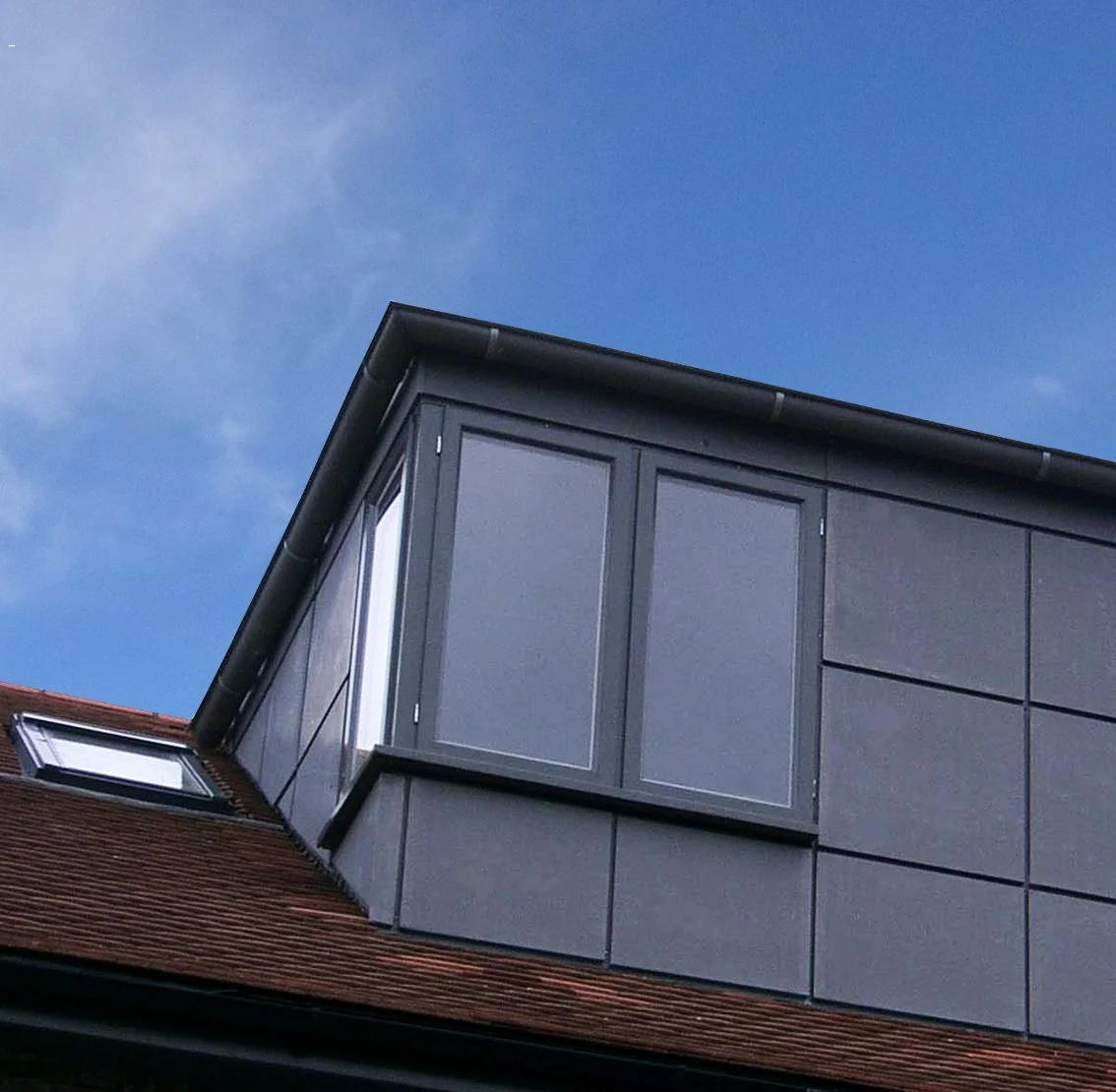 Close-up of a modern dormer window structure on a sloped roof, with a small skylight window and dark gray panels, under a partly cloudy sky.