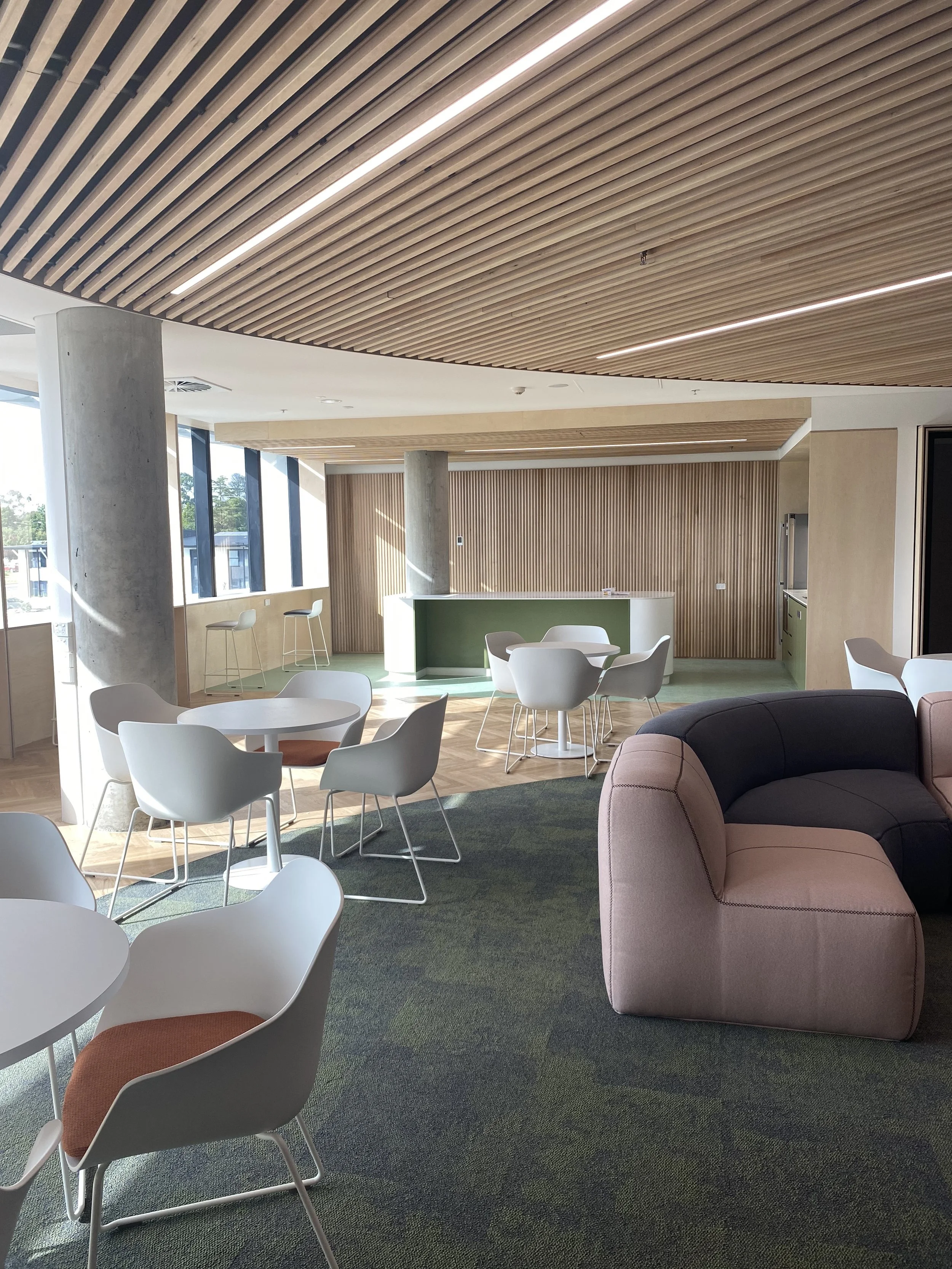 Modern office or lounge area with round white tables, white chairs with orange cushions, a pink armchair, a green reception desk, wooden slatted ceiling, large windows letting in natural light, and a green carpet.