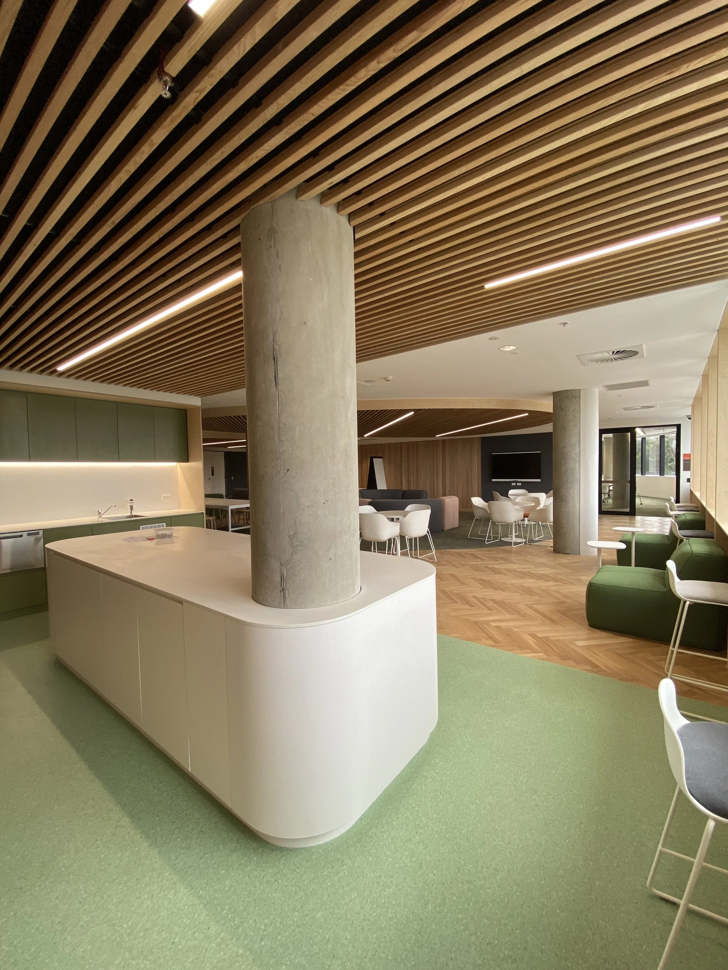 Modern interior with green carpet, white and green kitchen area, concrete columns, and seating area with chairs and tables under a wooden slatted ceiling with linear lighting.