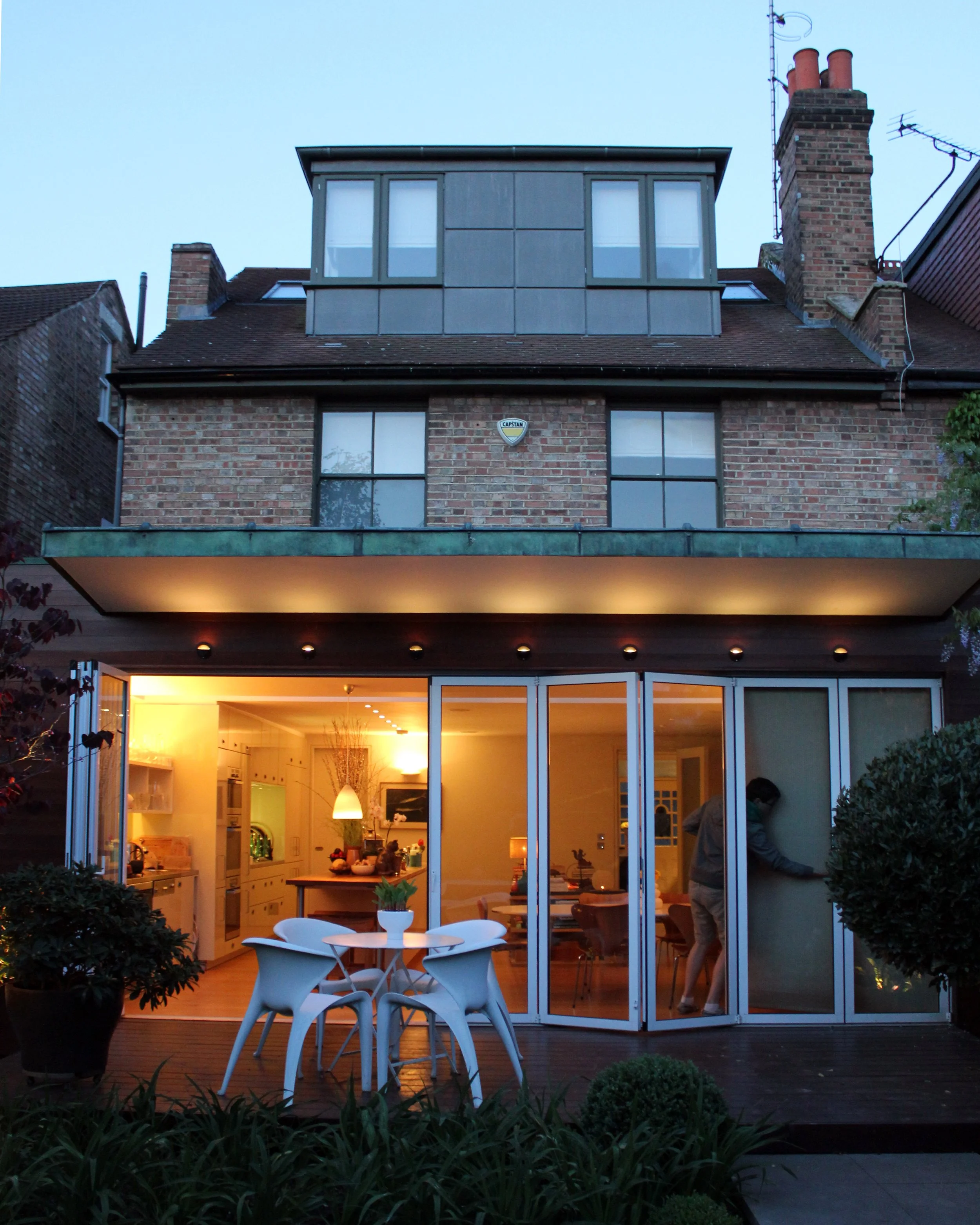 Nighttime view of a modern house with large glass sliding doors open to a well-lit interior. Outside, there is a small white table with four white chairs and surrounding greenery on a wooden deck.