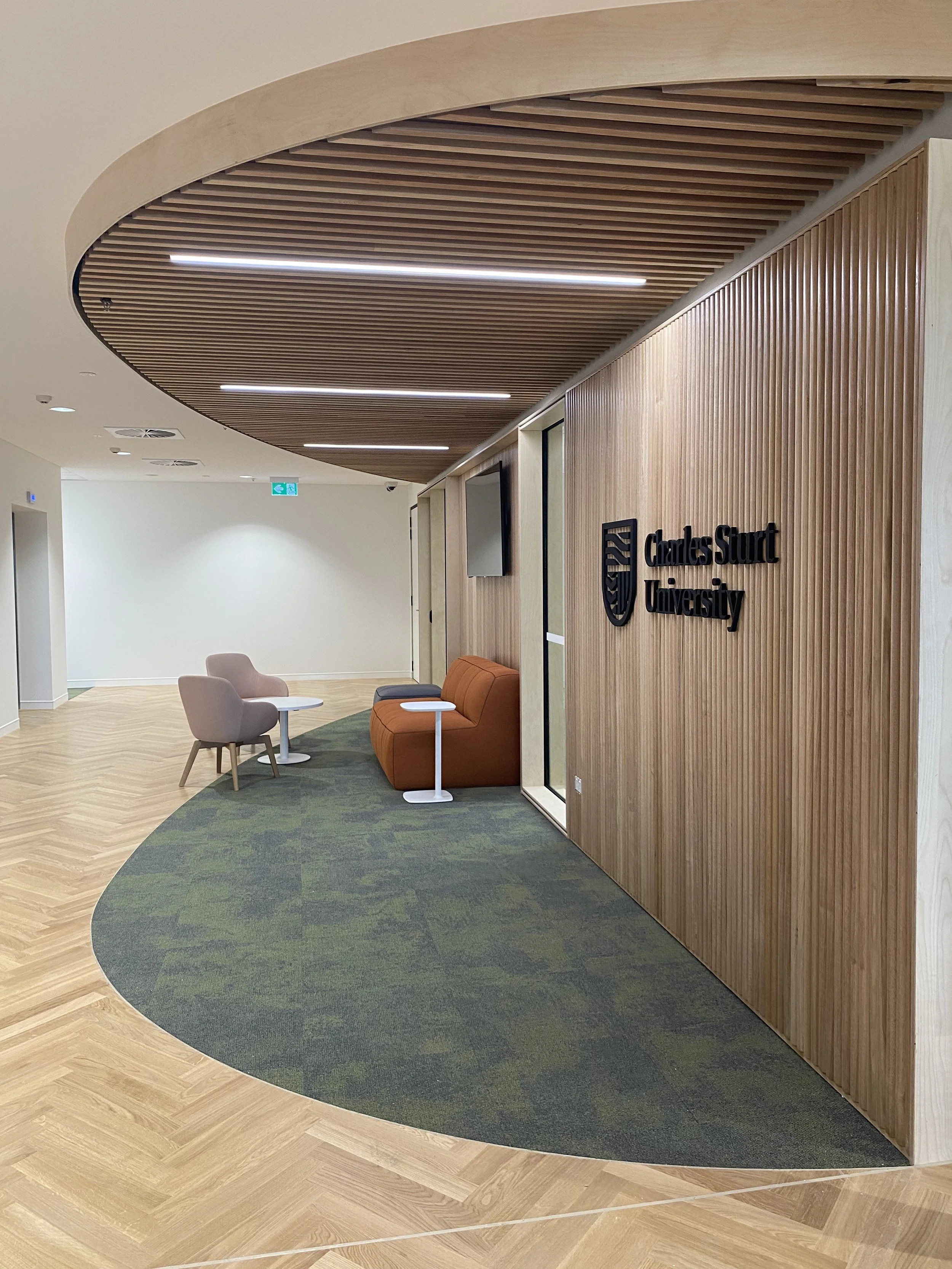 Interior view of a modern space at Charles Sturt University with wood paneling, a view of chairs and small tables, and a mix of light wood and green carpet flooring.