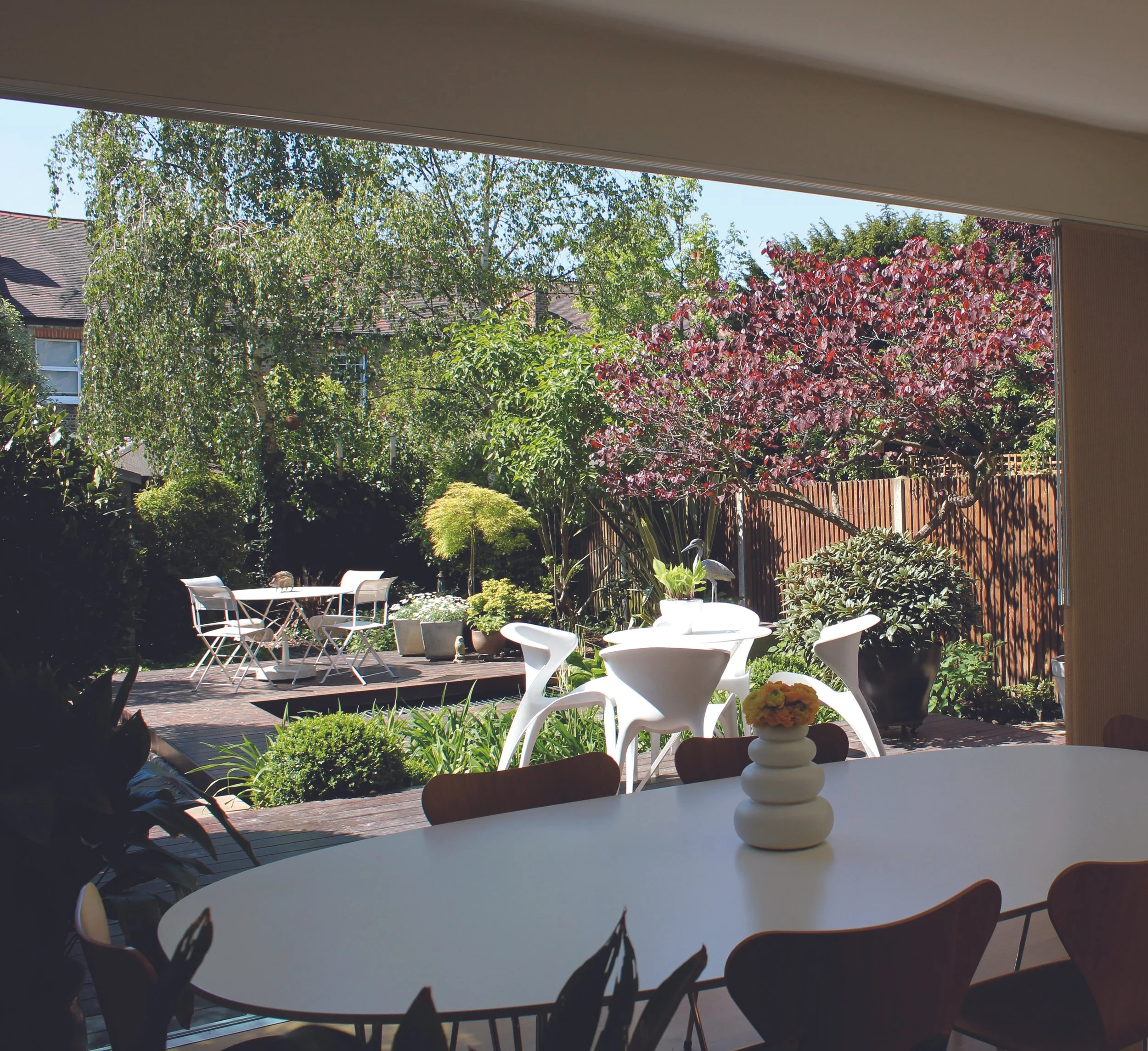 View of a backyard patio area with white round tables and chairs, potted plants, and lush green trees with some red foliage, seen from inside a house through a large open window.