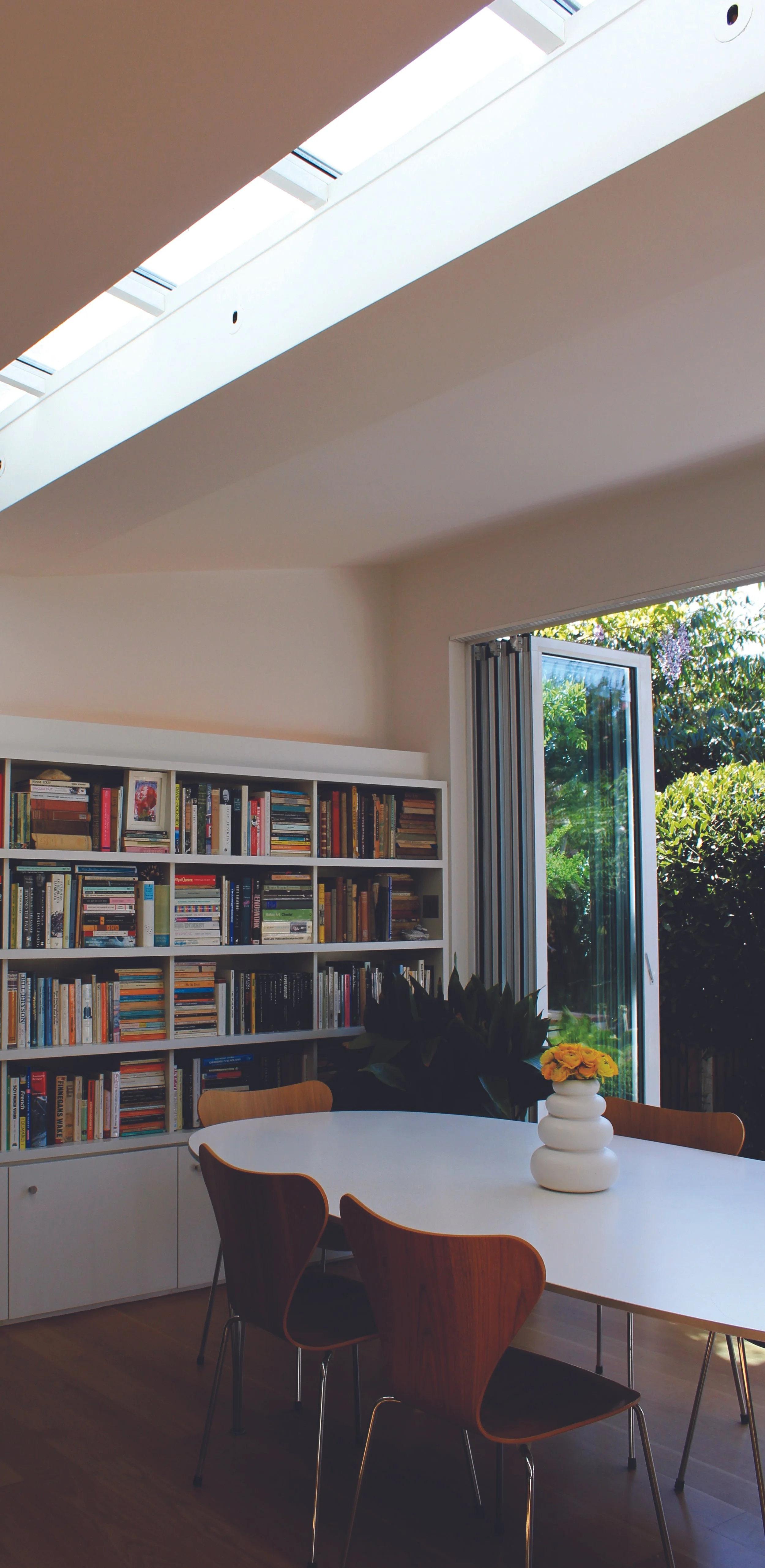 A well-lit room with a white bookshelf filled with books, a white table with a white vase holding yellow flowers, and wooden chairs. A large window with an open glass door reveals greenery outside, and sunlight streams into the room.