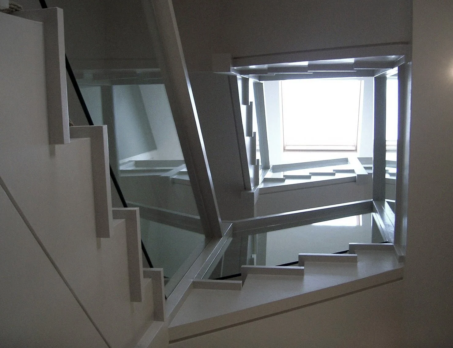 A spiral staircase viewed from below, with glass panels on the side and a large rectangular window at the top, allowing light to illuminate the staircase.
