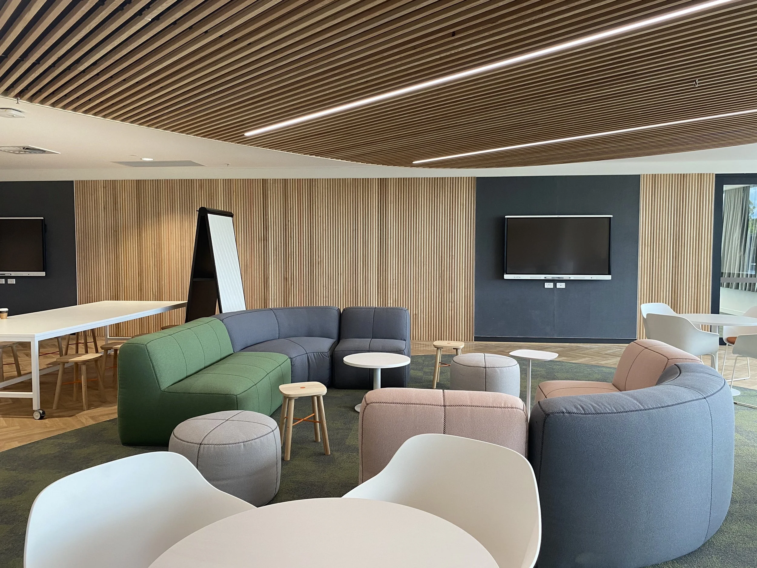 Modern open room with seating area, featuring a gray sofa, green and pink cushioned seats, white tables, large TV screens on the wall, and a wooden slatted ceiling with LED lighting.