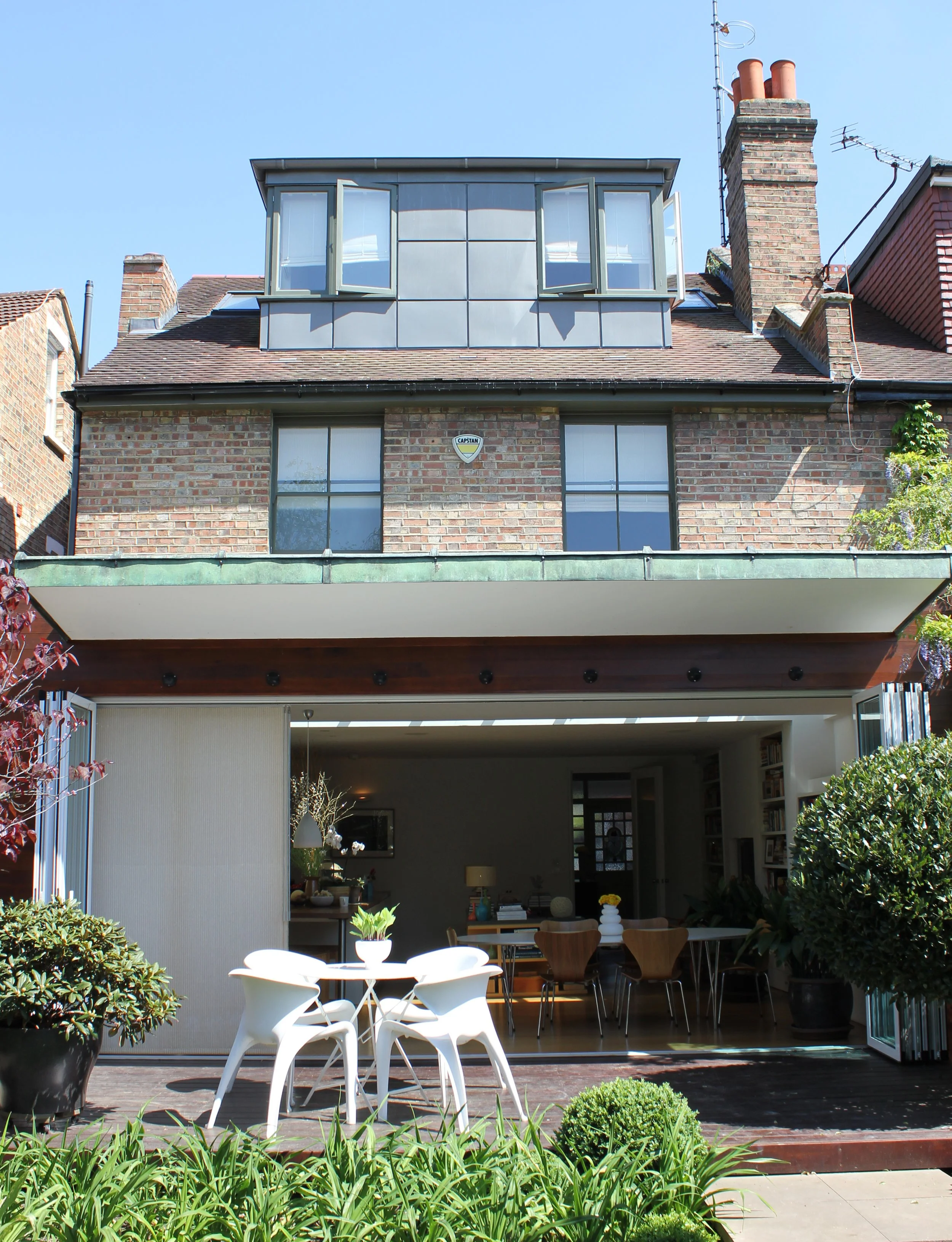 A modern residential building with an outdoor patio area, featuring white and wooden chairs, potted plants, and lush greenery, with large windows and a brick chimney.
