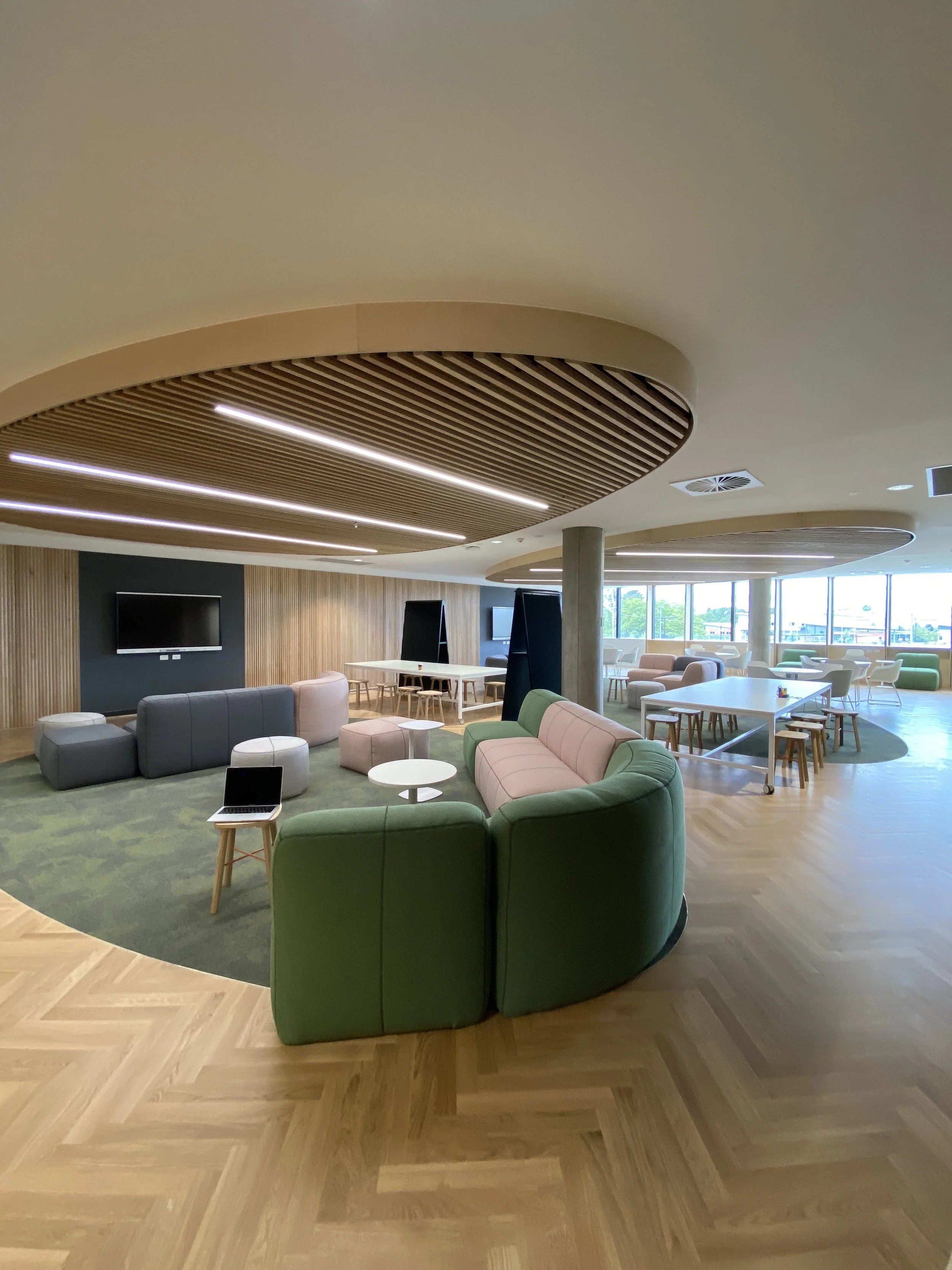 Modern lounge area with colorful sofas, round tables, and a wooden floor. Large windows on the right side let in natural light.