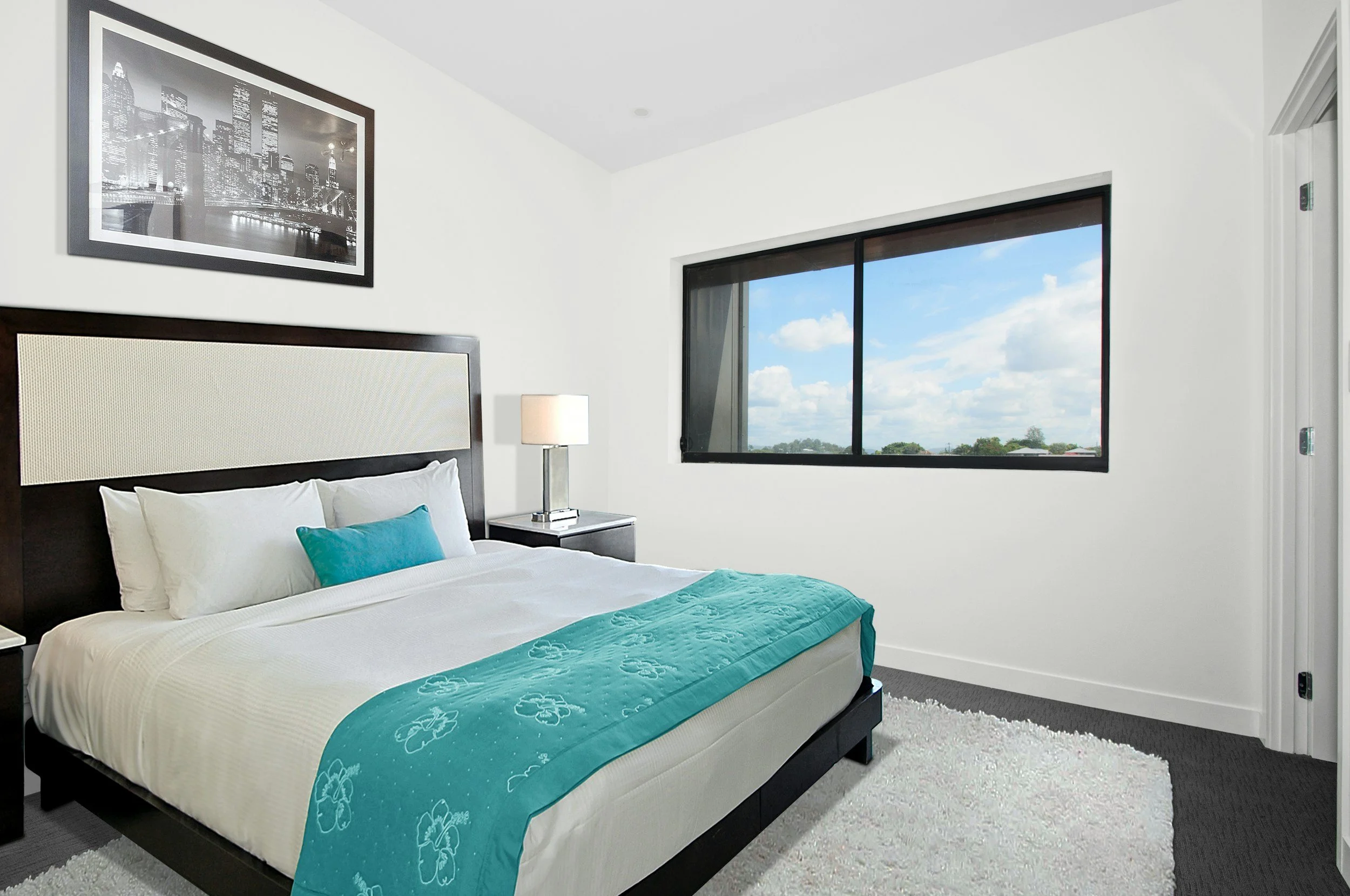 Habitación moderna con cama, cuadros en la pared, lámpara de mesa, ventana con vista a un cielo azul con nubes, alfombra blanca y paredes blancas.