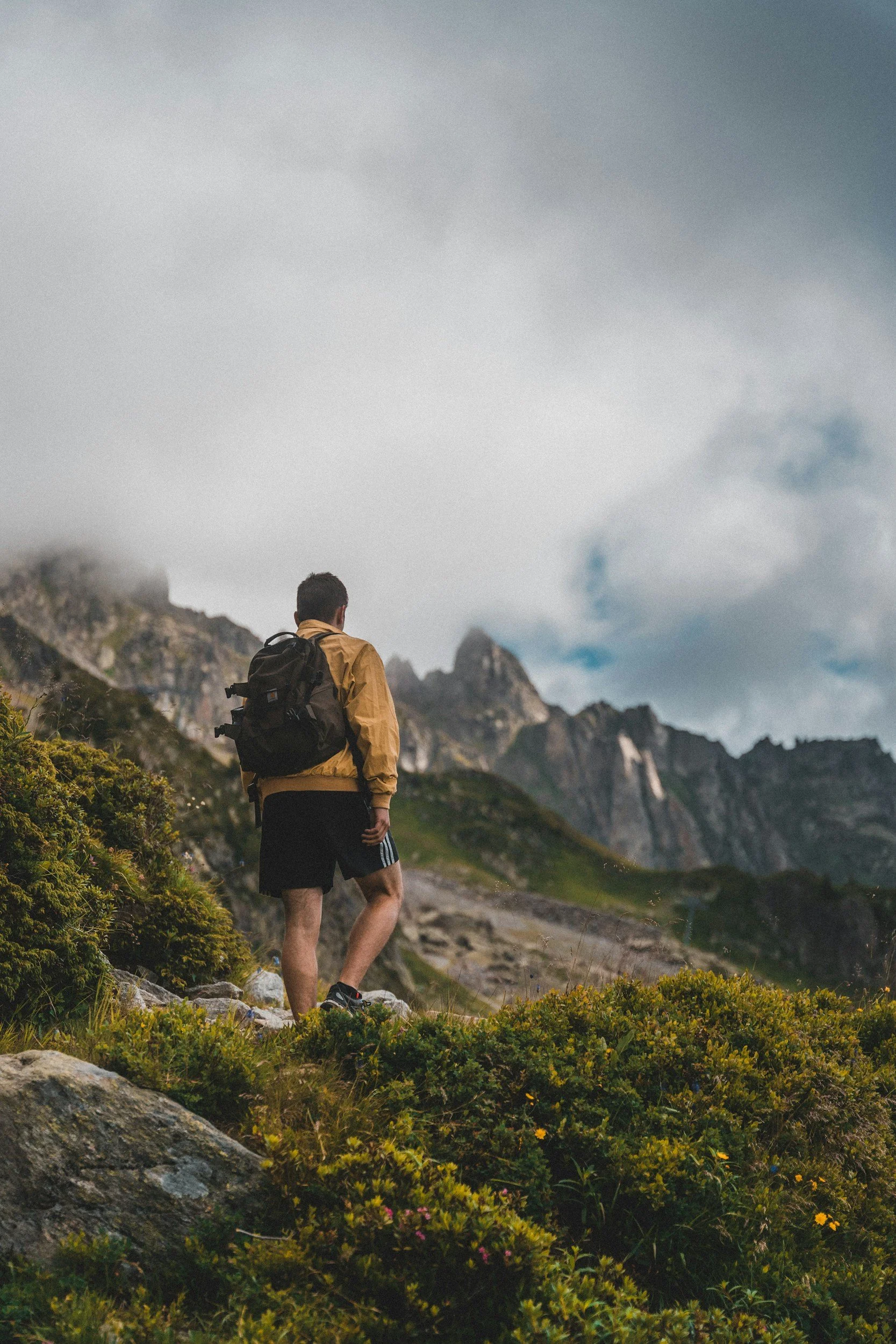 Persona con mochila explorando un paisaje montañoso con vegetación y formaciones rocosas, bajo un cielo nublado.