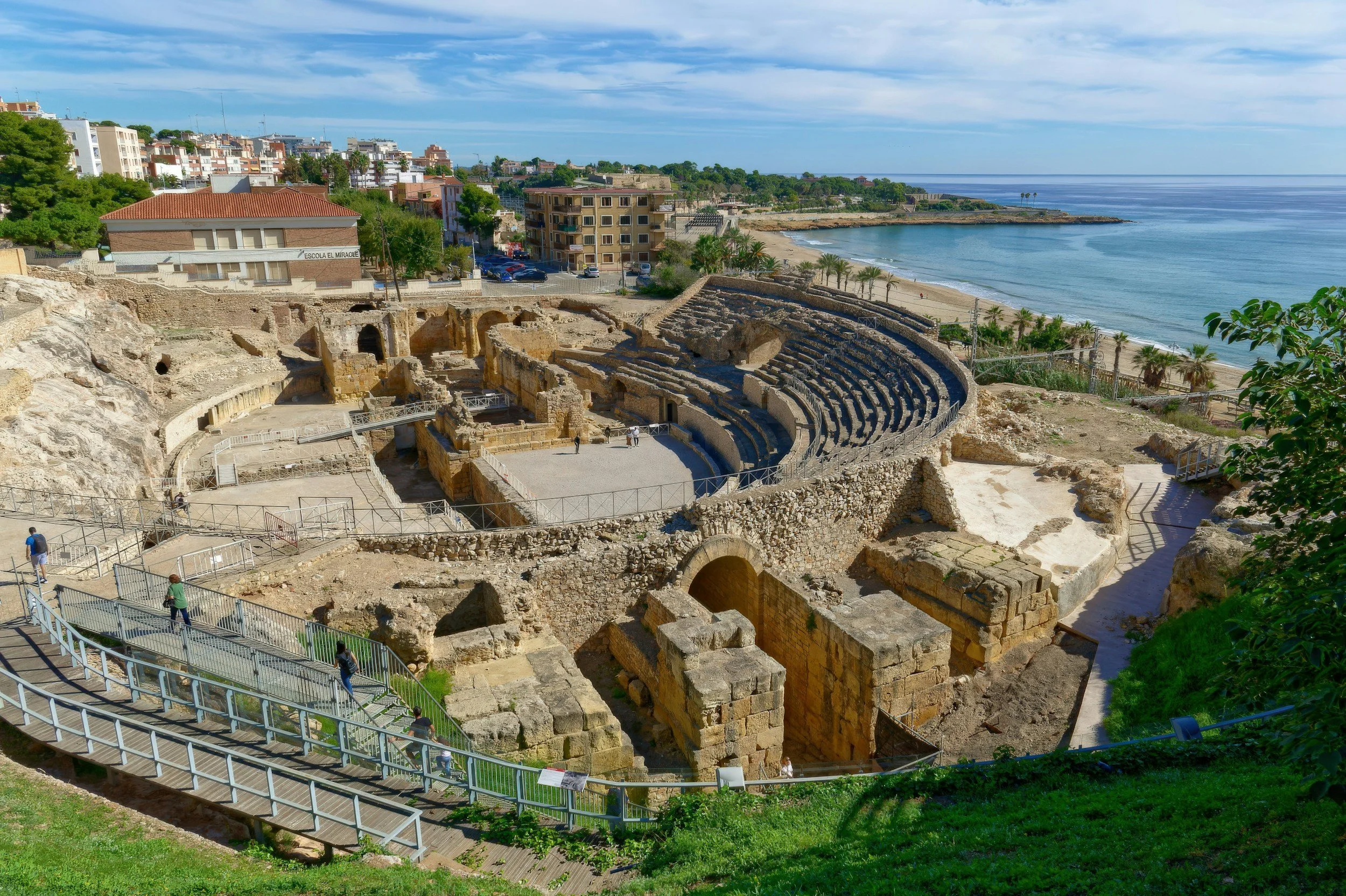 Vista del teatro romano en una zona costera, con edificios modernos, árboles y el mar en el fondo.