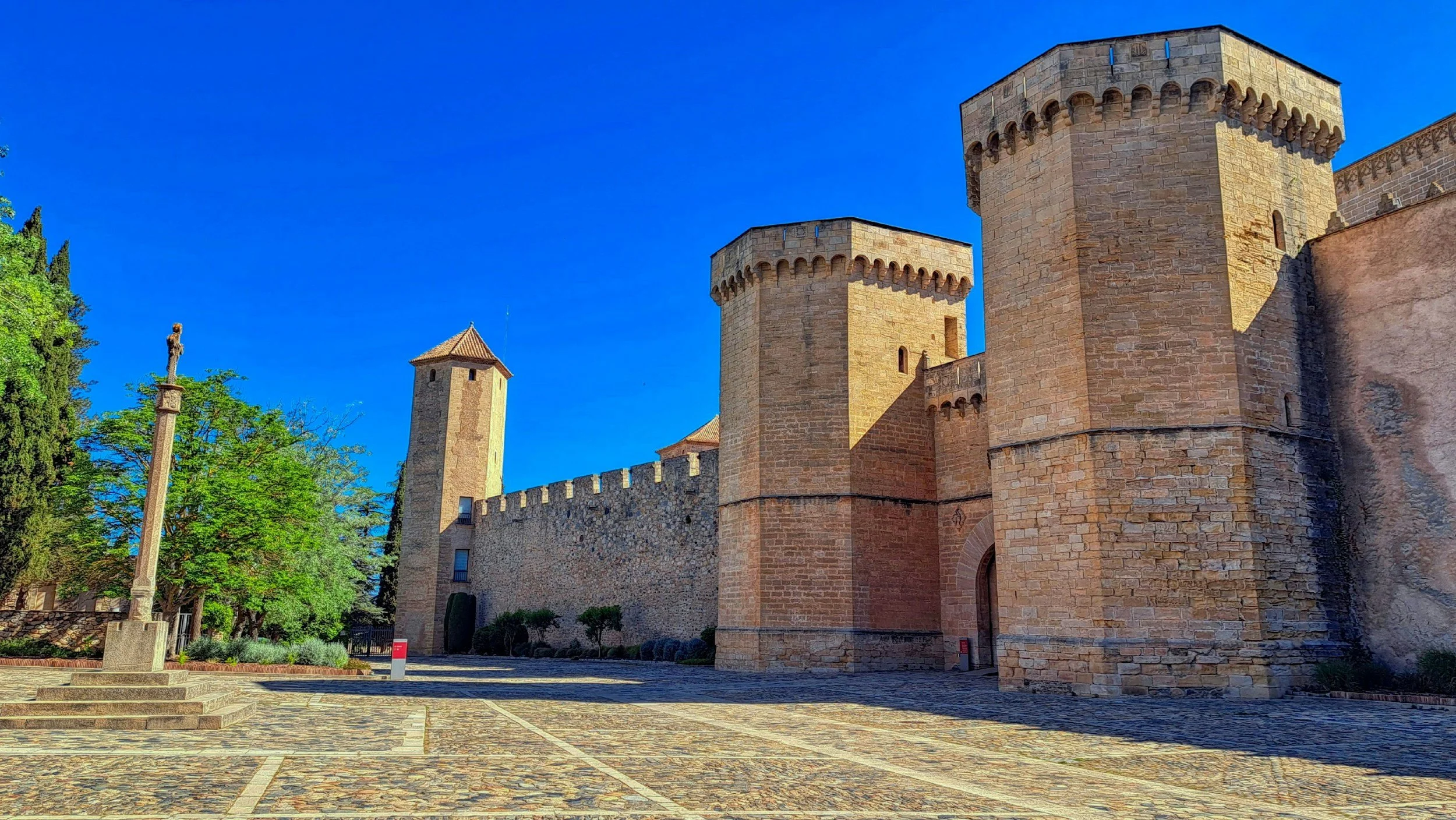 Castillo medieval de piedra con torres y muros, rodeado de árboles y un cielo azul claro.