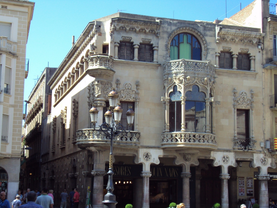 Edificio histórico con arquitectura elaborada y balcones ornamentados, en una calle con personas caminando y un poste de luz en primer plano.