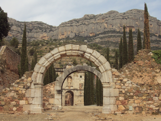 Antiguo arco de piedra en ruinas, con montañas y árboles en el fondo.
