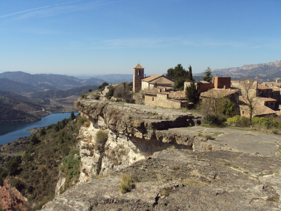 Vista de un pueblo antiguo con iglesia en una cima de roca, rodeado de paisaje montañoso y un río en el fondo.