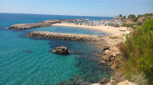 Vista de una playa con agua azul, rocas y un área con arena y sombrillas en el fondo.