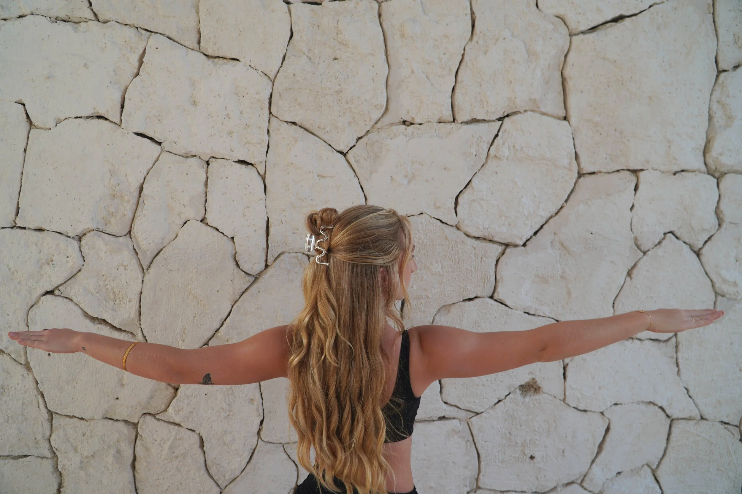 A woman with long blonde hair practicing yoga outdoors, standing with arms extended to the sides and palms facing down, in front of a stone wall.