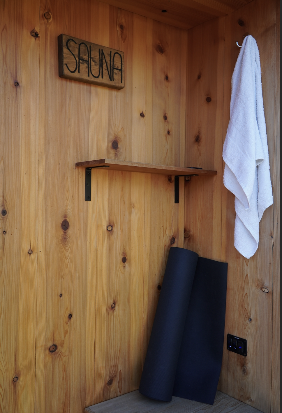 Wood-paneled wall with a wooden 'Sauna' sign, a towel hanging on a hook, and two rolled-up yoga mats leaning against the wall.
