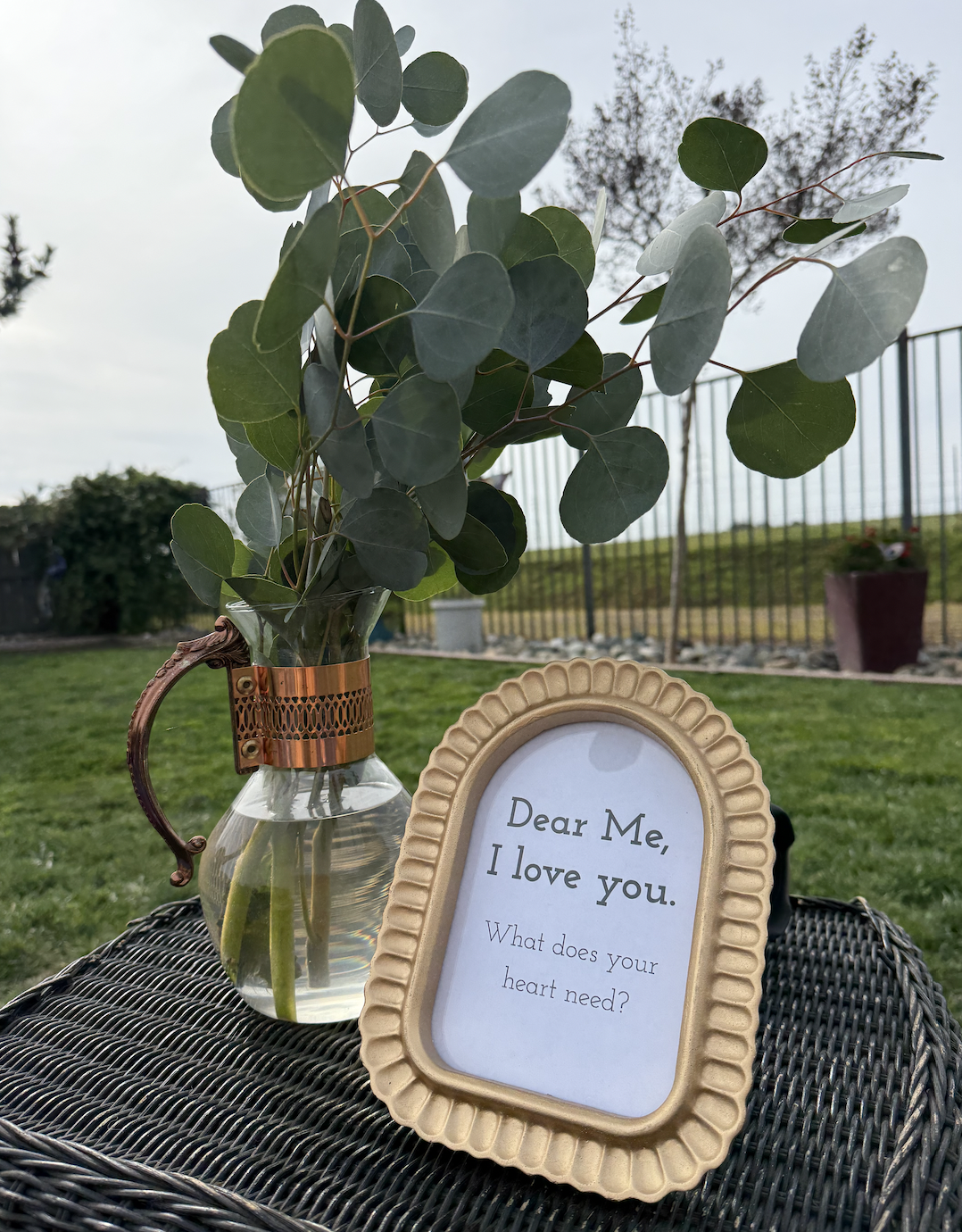 A small glass vase with eucalyptus leaves and a framed sign that reads "Dear Me, I love you. What does your heart need?" on a woven surface outdoors with grass, trees, and a fence in the background.
