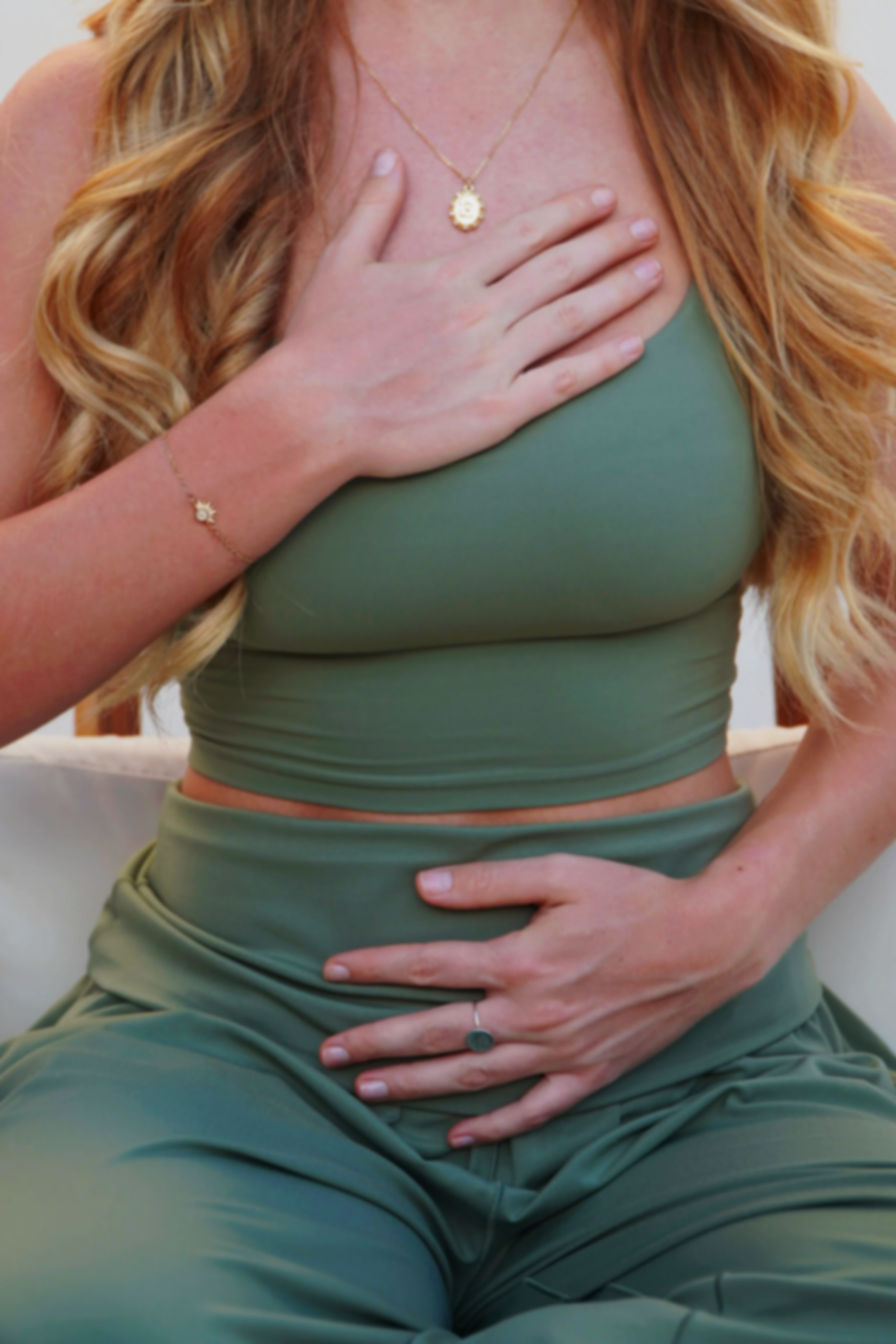 Close-up of a woman with long blonde hair wearing a green top and matching leggings, sitting cross-legged with one hand on her chest and the other on her stomach, wearing jewelry including a necklace, bracelet, and ring, in a sitting position.