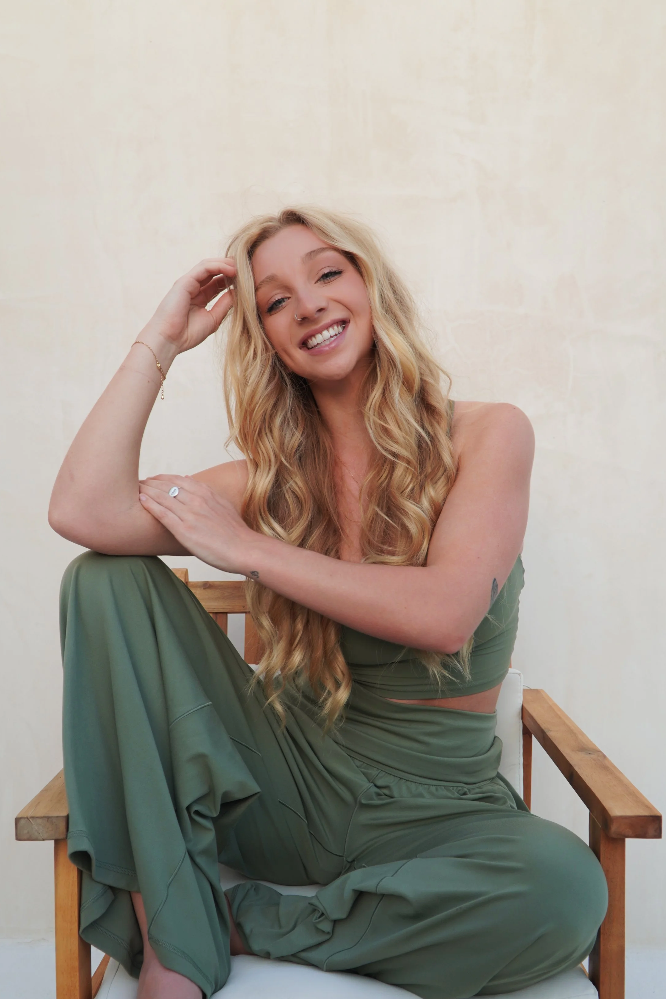 A smiling woman with long curly blonde hair, wearing a nose ring and a gold bracelet, sitting on a wooden chair with one leg crossed over the other, in front of a plain light-colored wall.