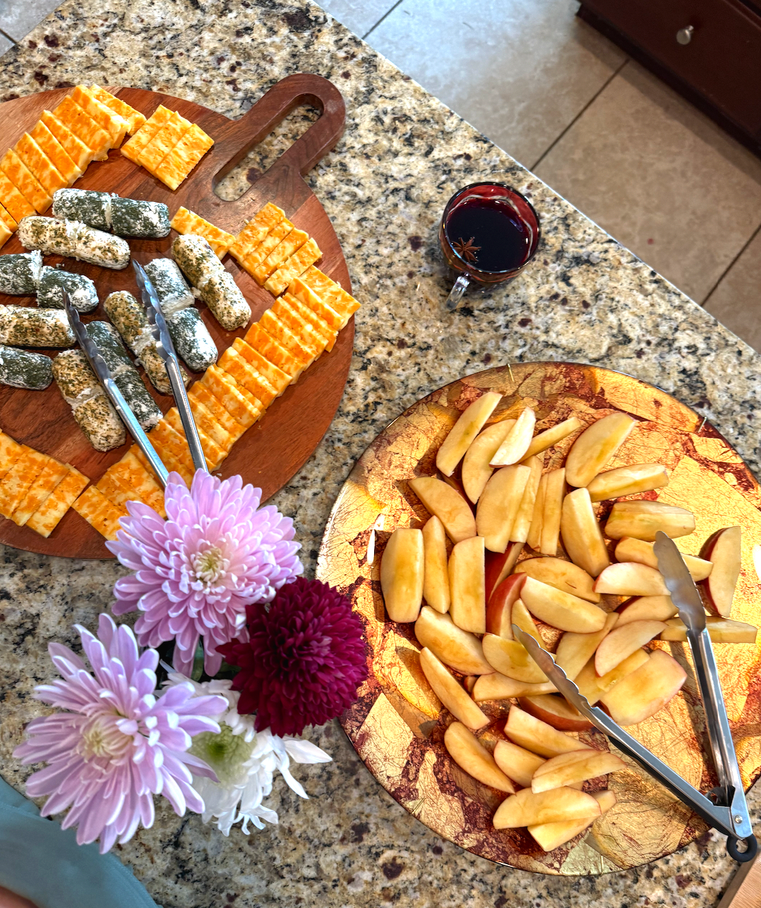 Variety of cheeses on a wooden cutting board, sliced apples on a decorative gold platter, a glass of red wine, and a vase with pink, white, and maroon flowers on a granite countertop.