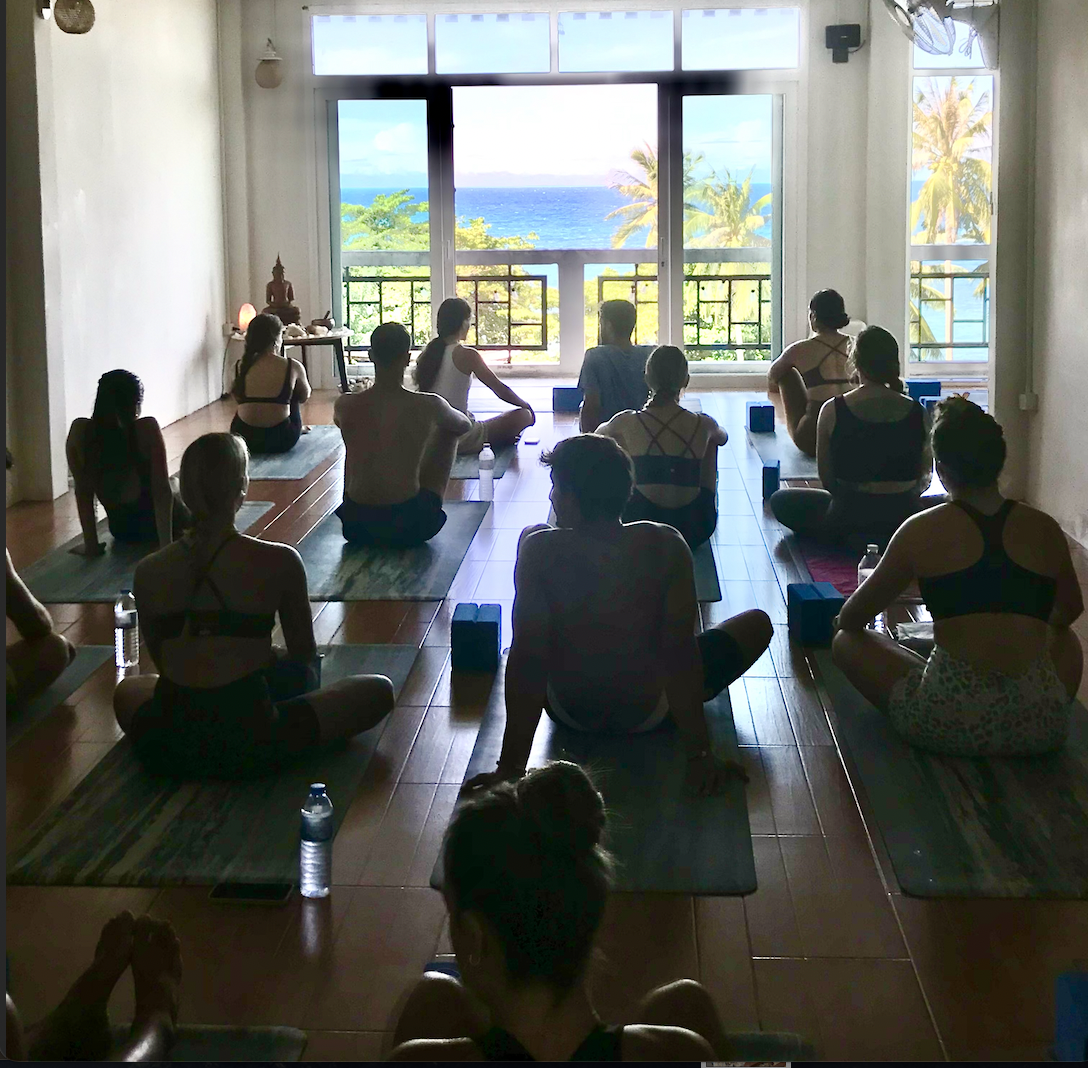 People participating in a yoga class in a room with a view of the ocean, palm trees, and blue sky through large open doors.