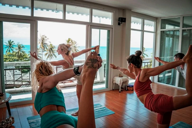 Group of women practicing yoga in a bright room with large windows overlooking a tropical beach, some in a backbend pose and one on a balcony stretching outdoors.