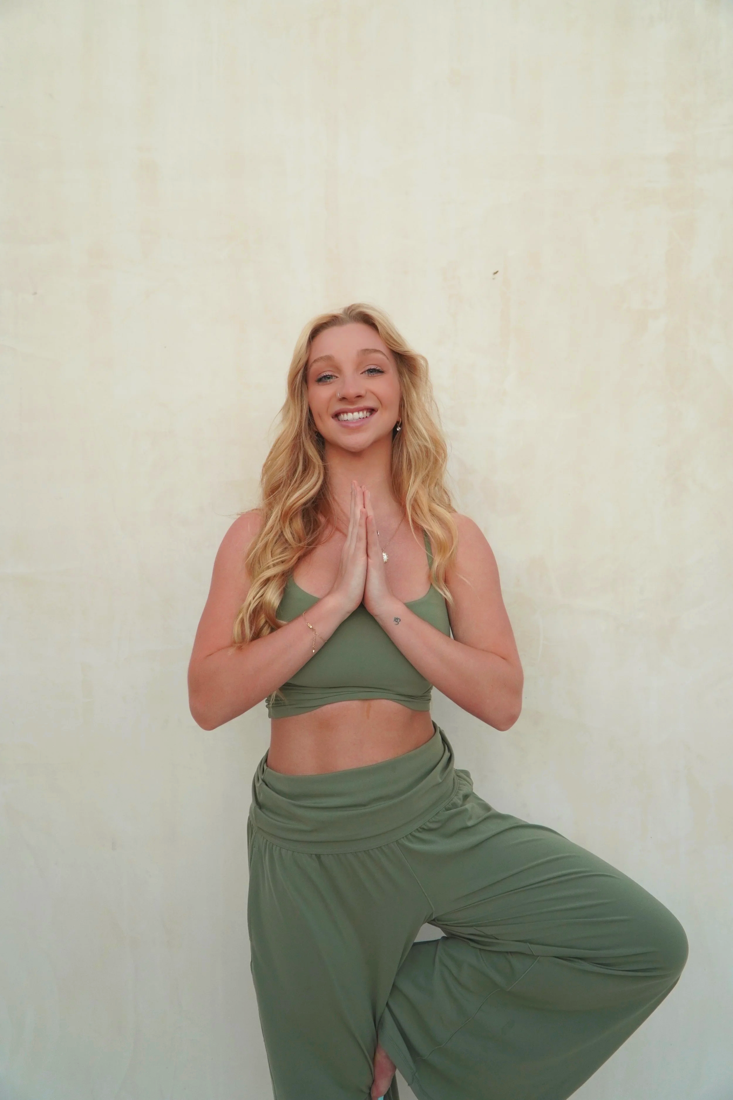 A young woman with long blonde hair posing in a yoga pose against a light yellow wall, wearing a green sports bra and matching loose pants, smiling with hands in prayer position.