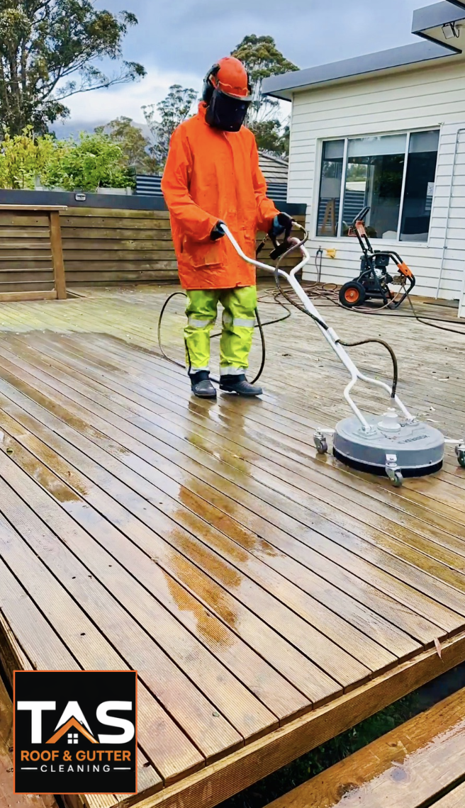 A specialist using pressure cleaning a wet wooden deck using a floor cleaning machine, wearing an orange raincoat, a red helmet, and green waterproof pants, with a house, trees, and cleaning equipment in the background.