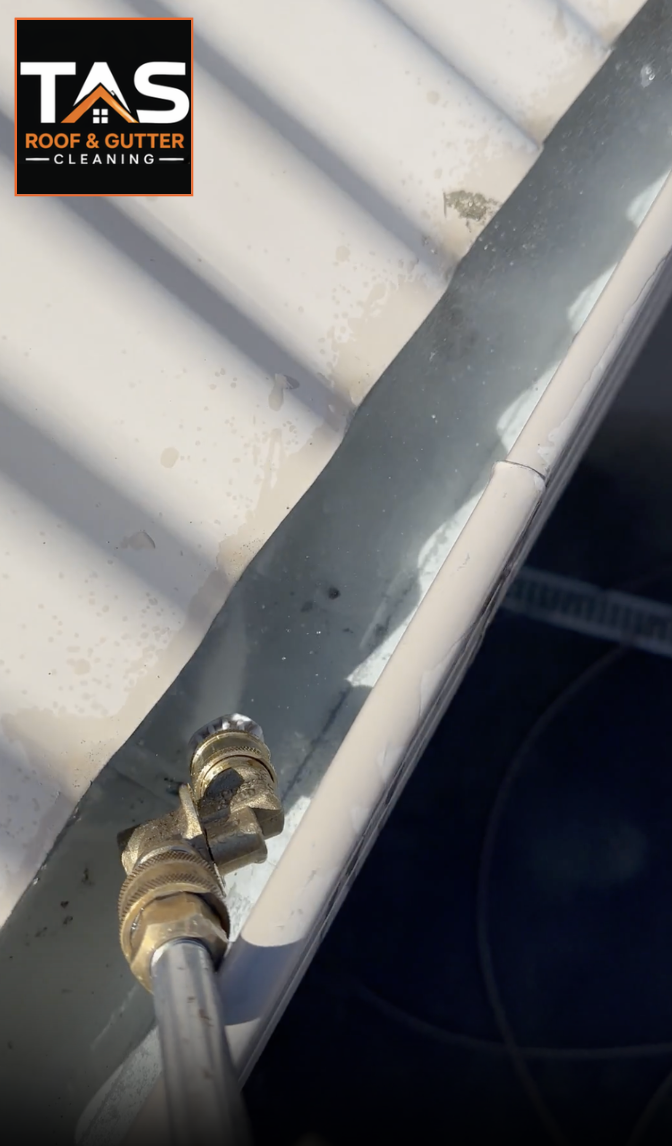Close-up of a metal roof gutter with a hose spraying water and the logo of TAS Roof & Gutter Cleaning