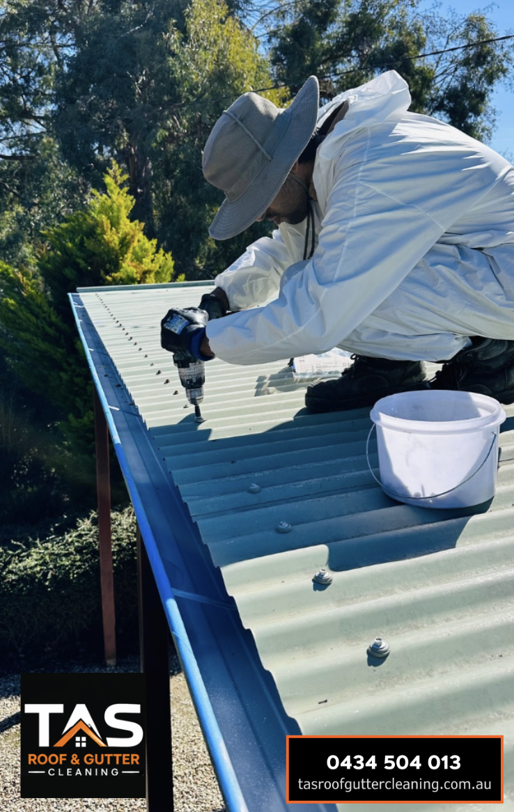 A person wearing a wide-brimmed hat and white protective clothing working on cleaning or repairing a corrugated metal roof with a power drill, with a bucket nearby, outdoors with trees in the background.