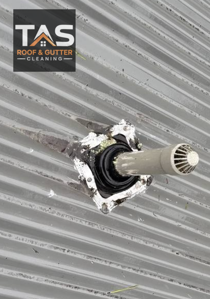 Capped roof pipe with surrounding flashing and dark surface staining along the corrugated metal roof.