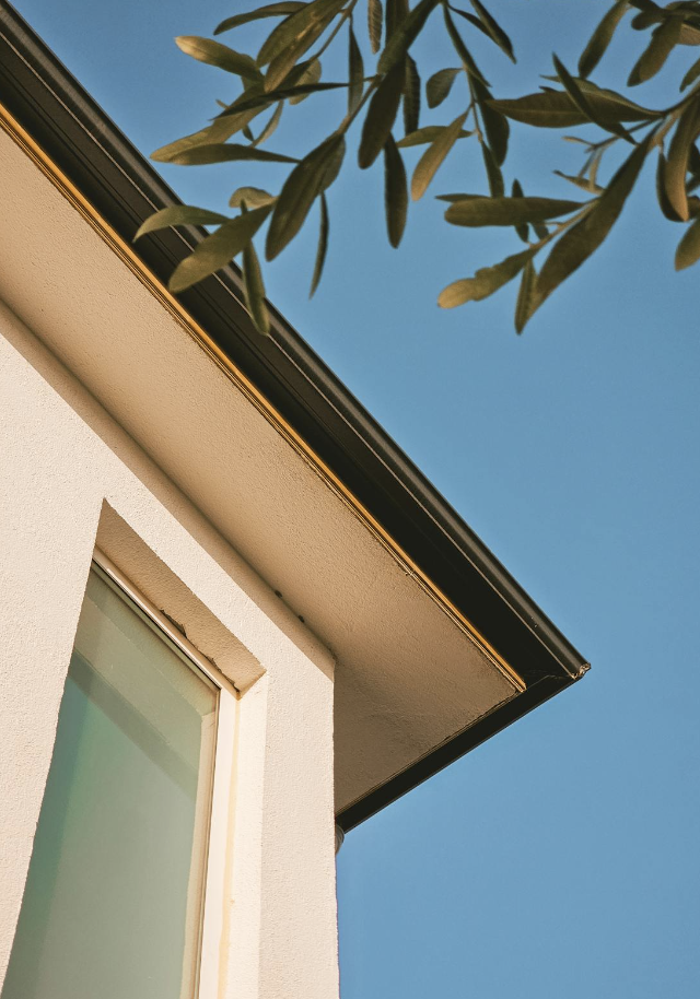 A close-up shot of a modern house with a beige stucco exterior, a large window, and a black gutter, with green leaves and blue sky in the background.