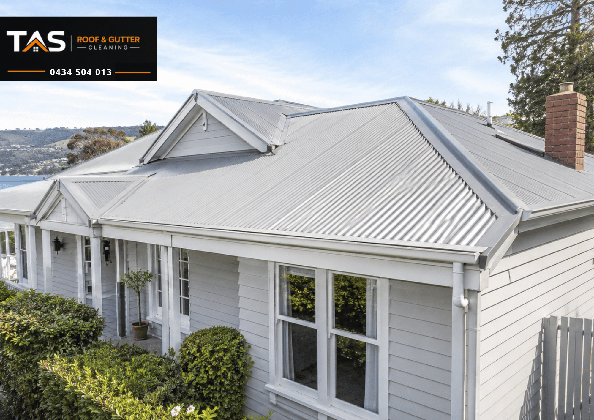 A white house with a corrugated metal roof. Roof repairs for house ready for selling in TAS. The house has large windows, a white porch, and brick chimney. There are lush bushes in front and a scenic lake and hills in the background.