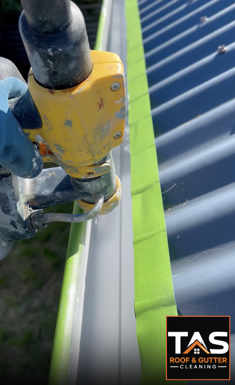 Applying a protective coating inside a metal gutter, with edges masked beside a corrugated roof, by TAS Roof & Gutter Cleaning.