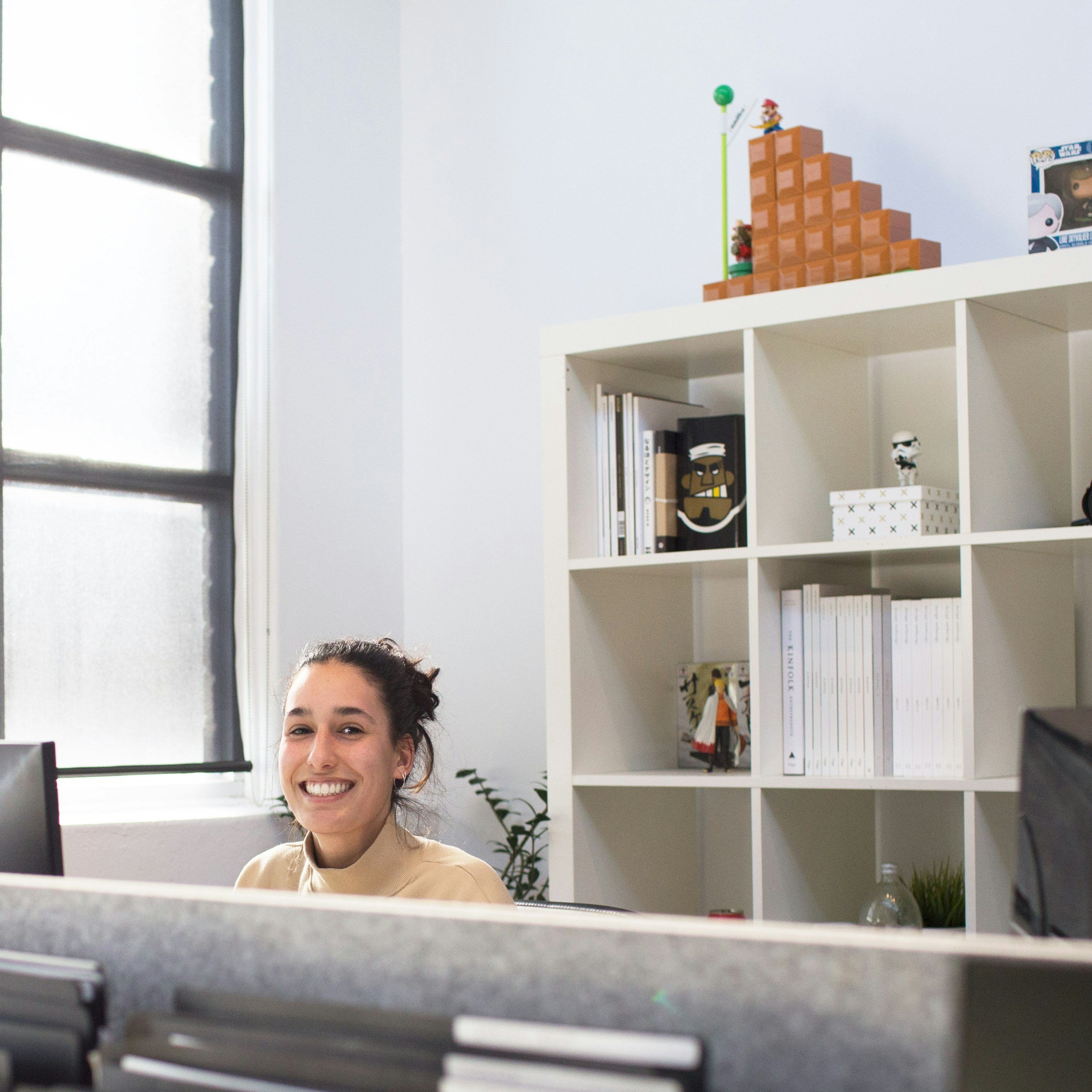 A young woman sitting at a computer desk, smiling, with a white bookshelf filled with books and decorations in the background. There is a window on the left side of the image.