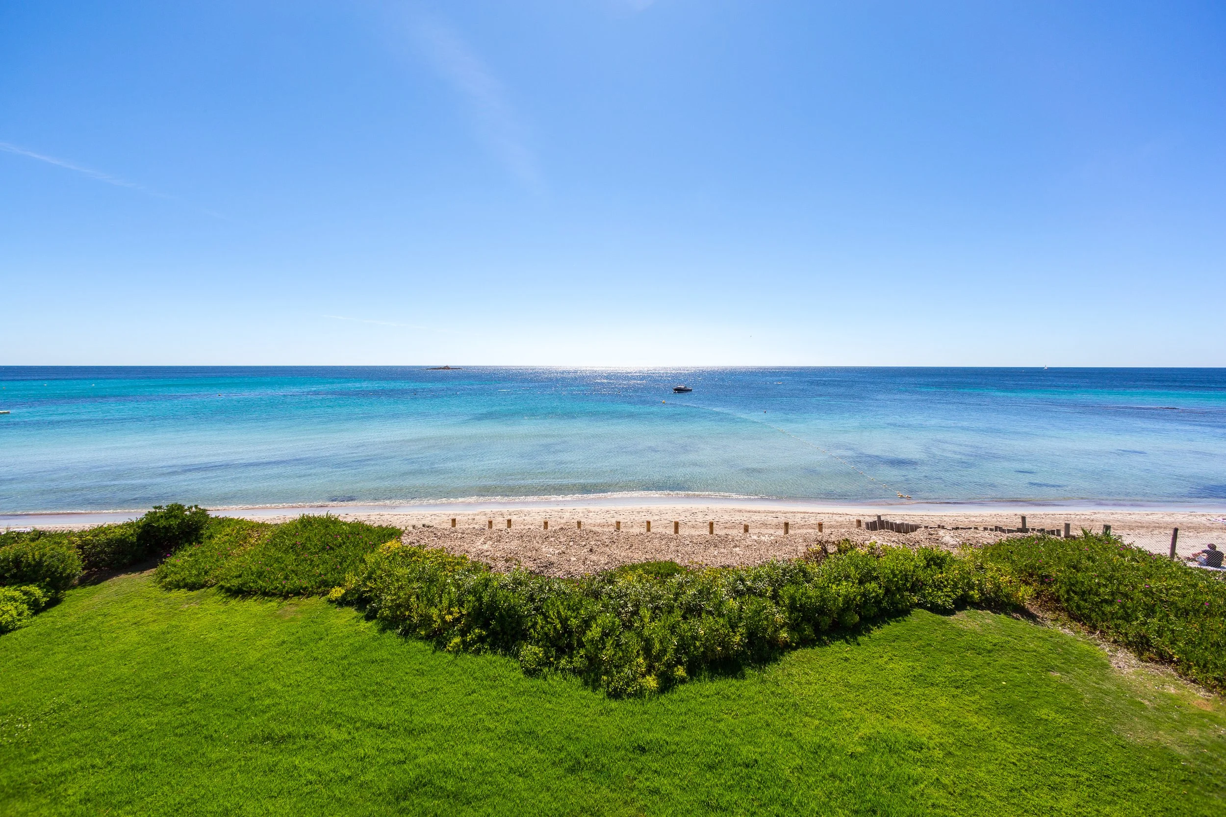 Plage avec eau bleue et ciel clair, vue depuis la verdure bordant le sable