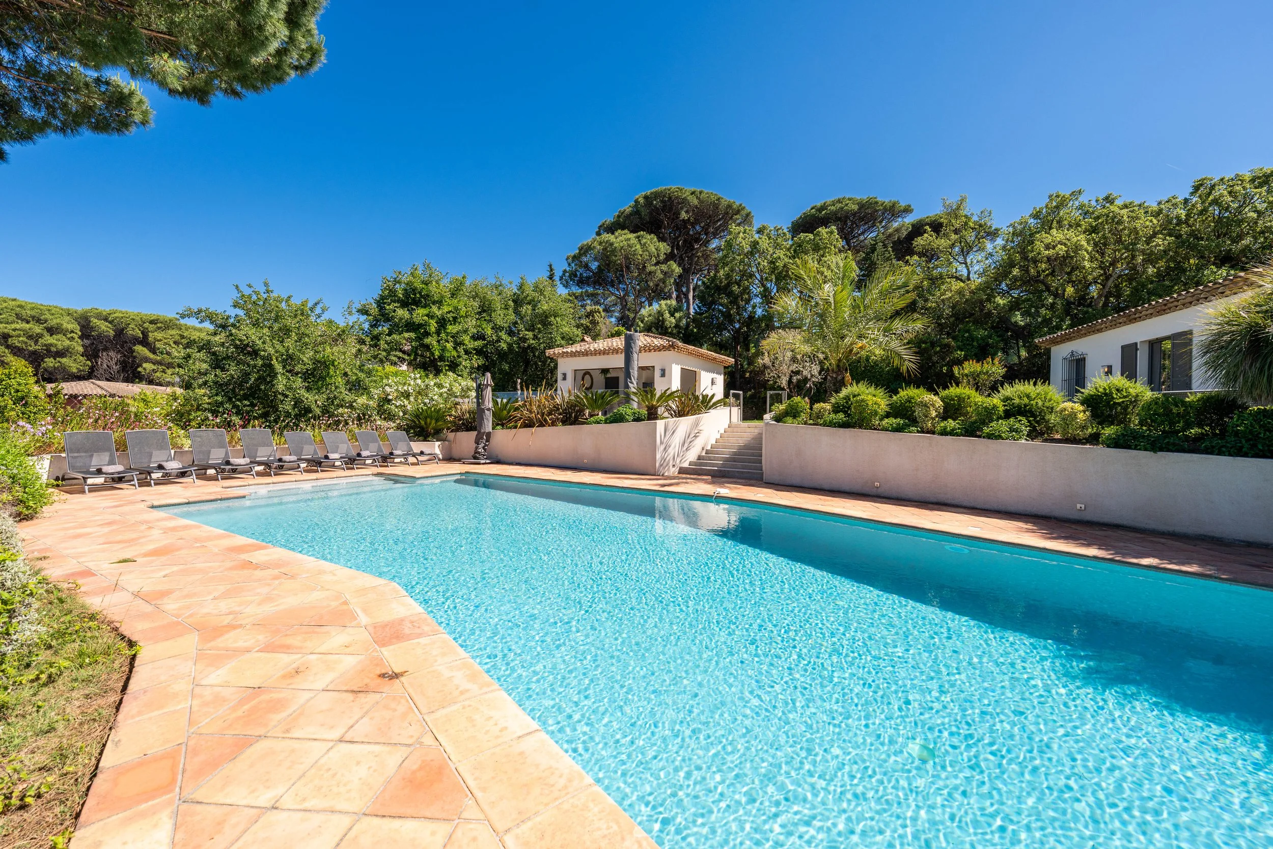 Piscine extérieure avec chaises longues, entourée de végétation et de maisons blanches, sous un ciel bleu clair.