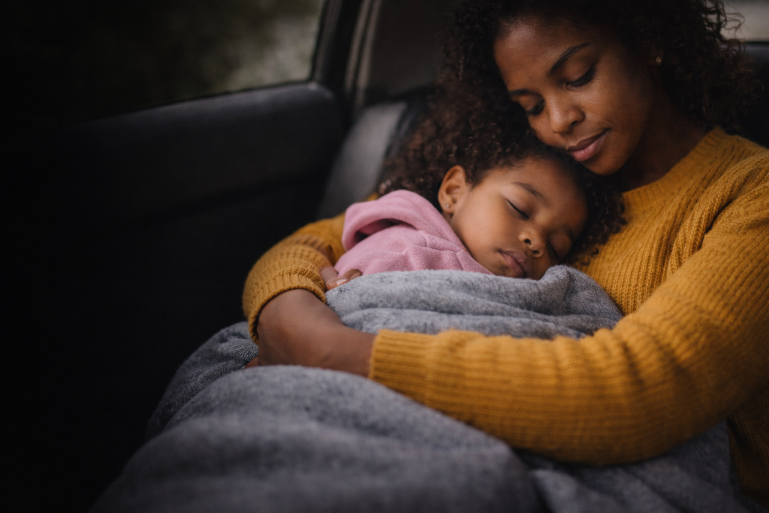 A woman with curly hair in a mustard yellow sweater hugging a young girl in a pink hoodie while sitting in a vehicle, both with closed eyes as they rest.
