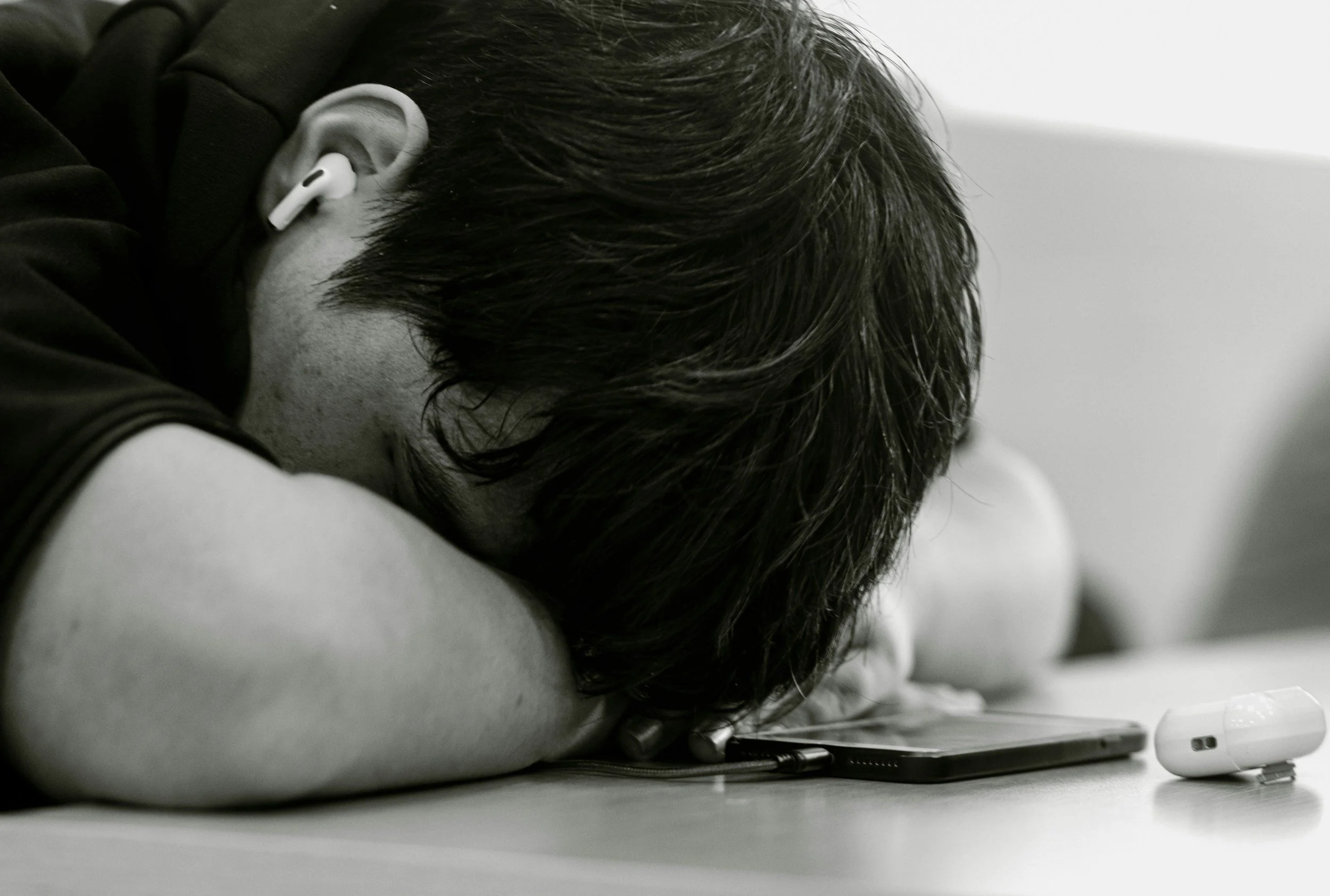 Person resting their forehead on a table, next to a tablet and wireless earbuds on the table.