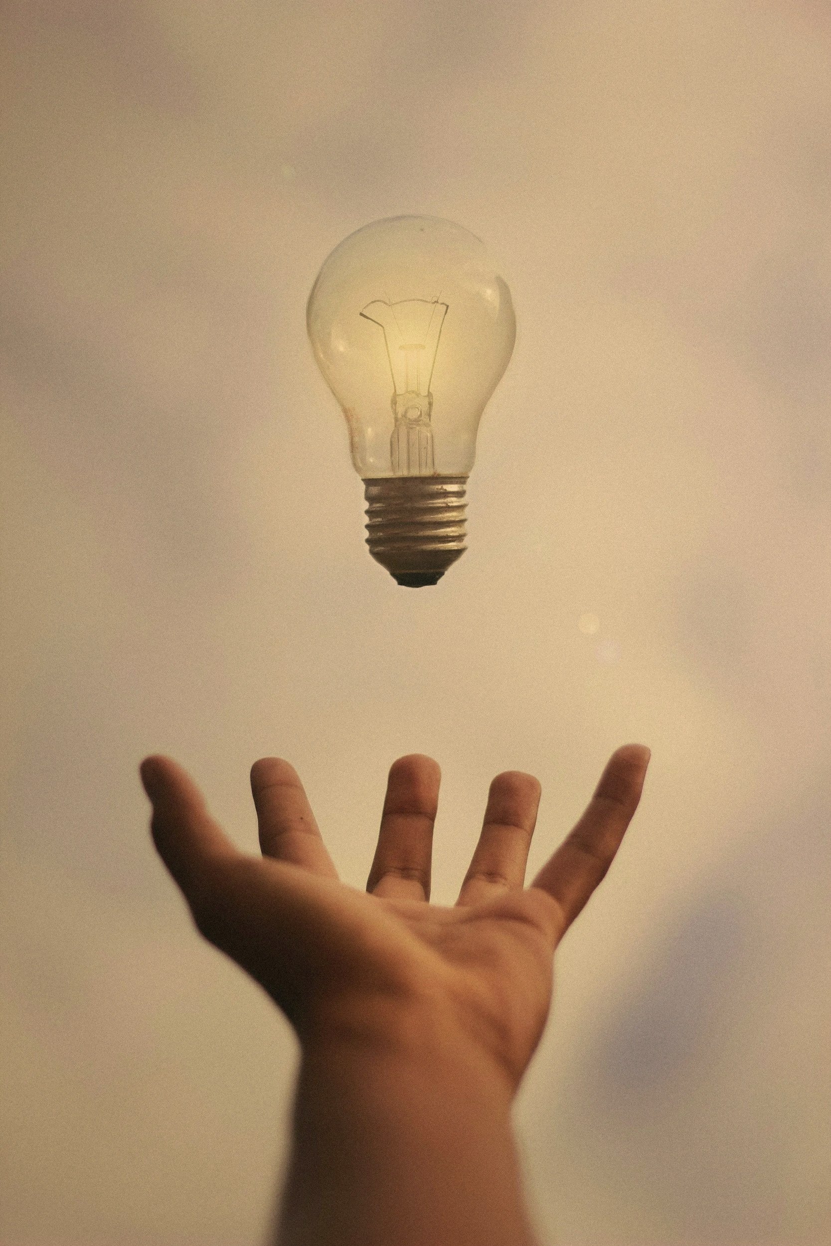 An outstretched hand with fingers reaching upwards toward a floating light bulb.