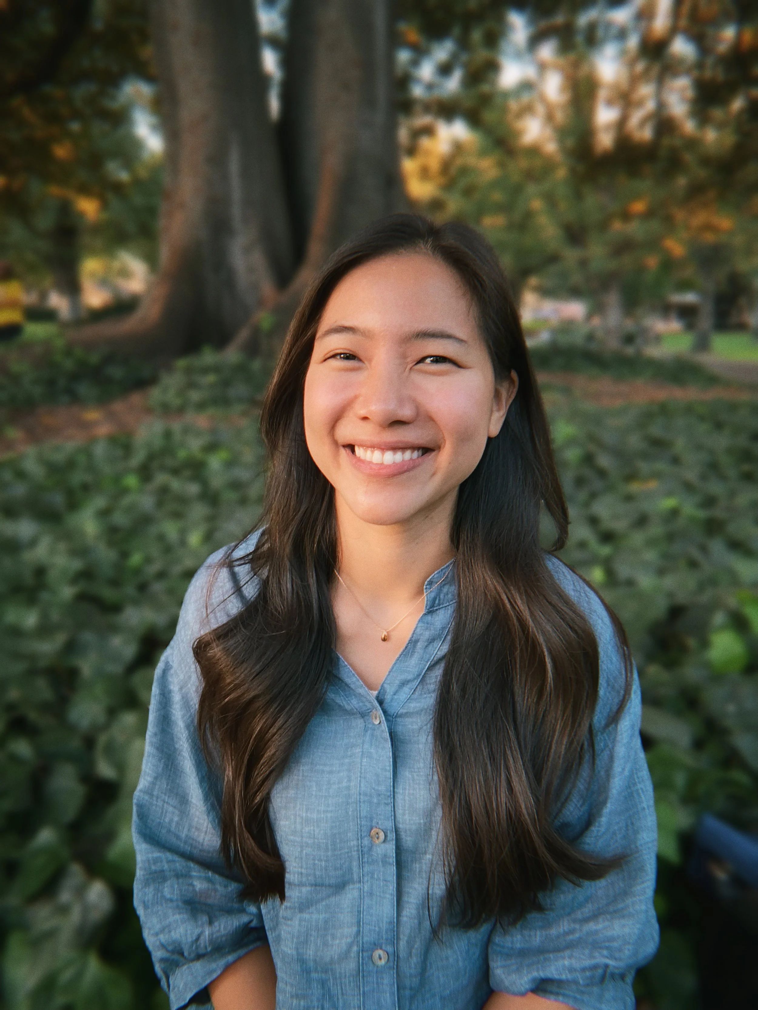 A young woman with long dark hair smiling outdoors in front of trees and greenery, wearing a light blue button-up shirt and a necklace.