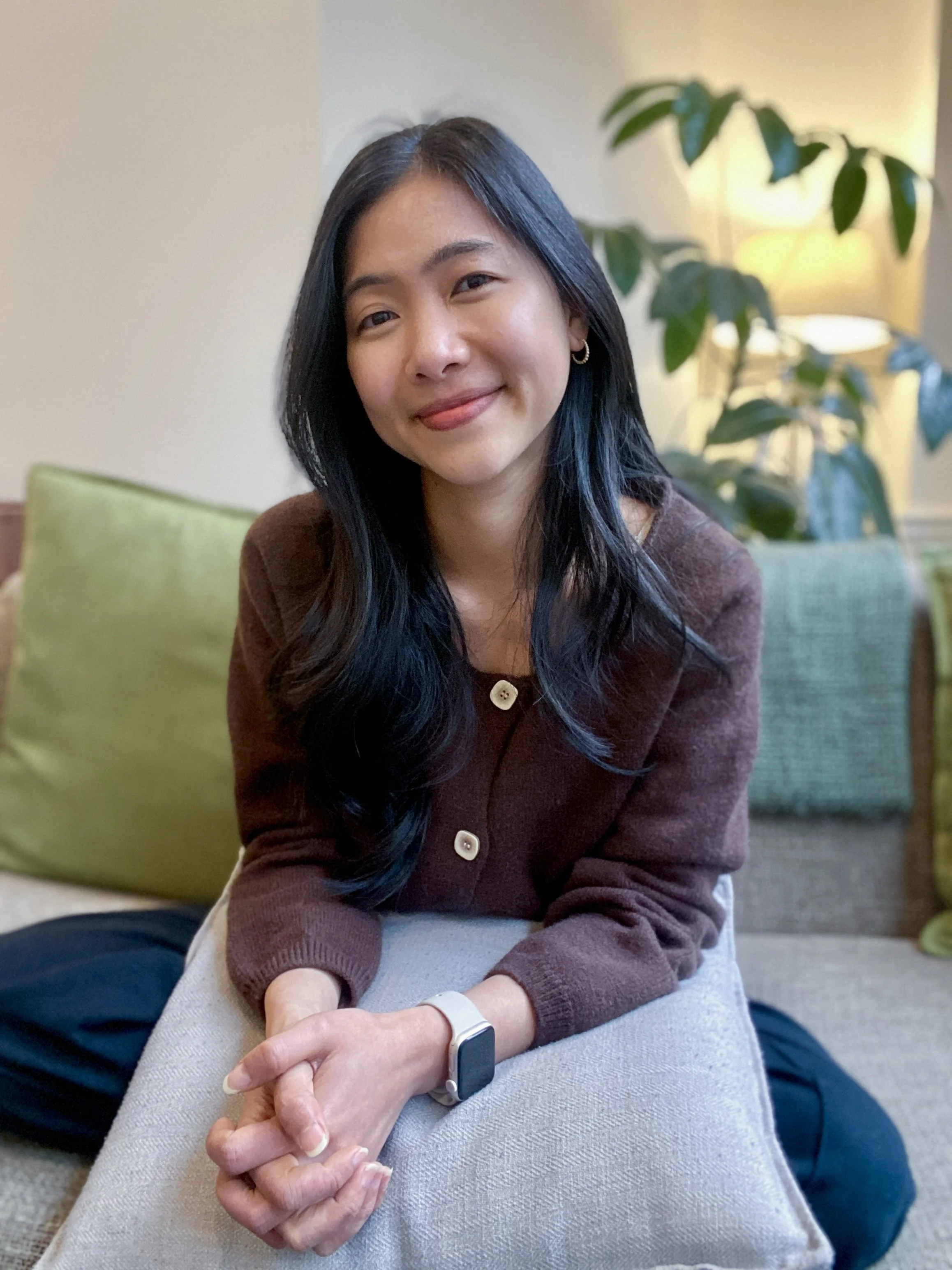 A woman with long dark hair and a warm smile, sitting on a sofa with a green pillow behind her, in a cozy living room with a plant and lamp in the background.