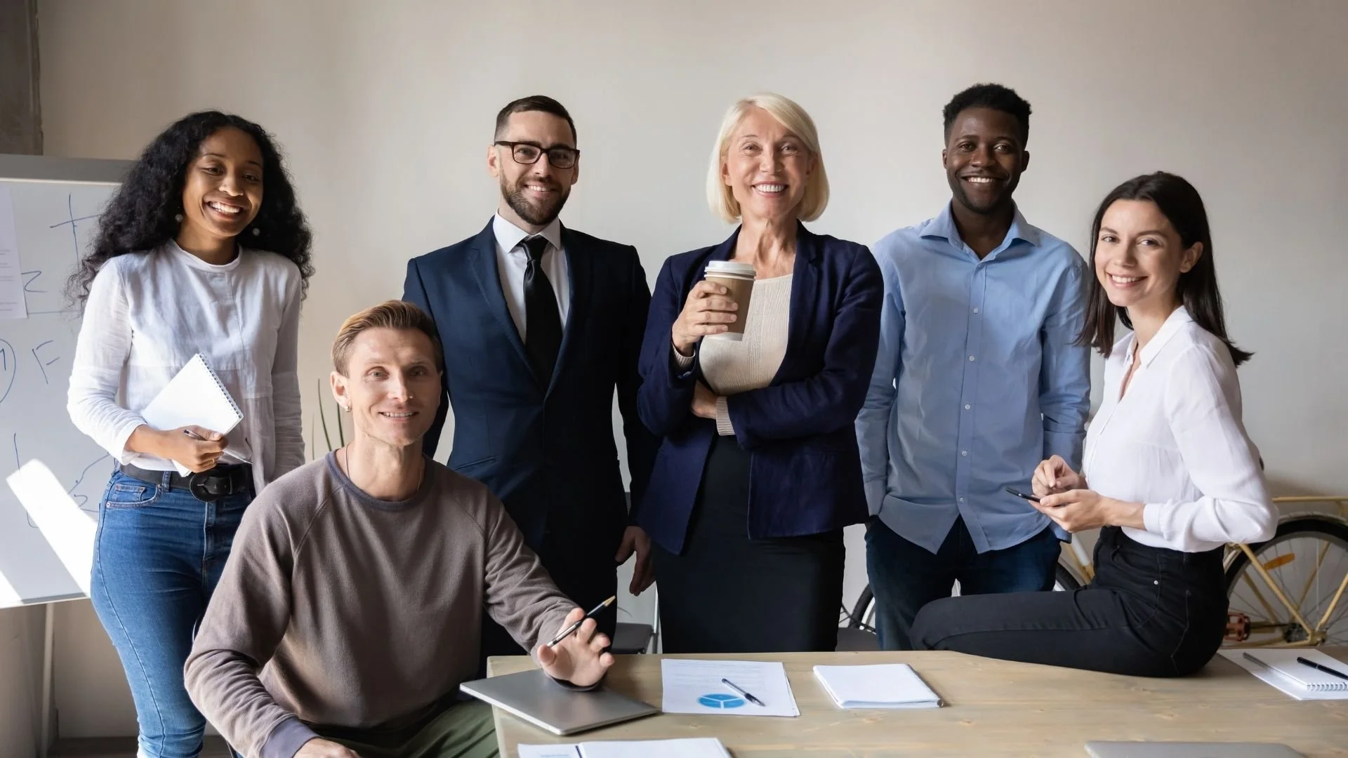 Group of six diverse professionals in a meeting or workshop, smiling and looking at the camera, with a whiteboard and bicycle in the background.