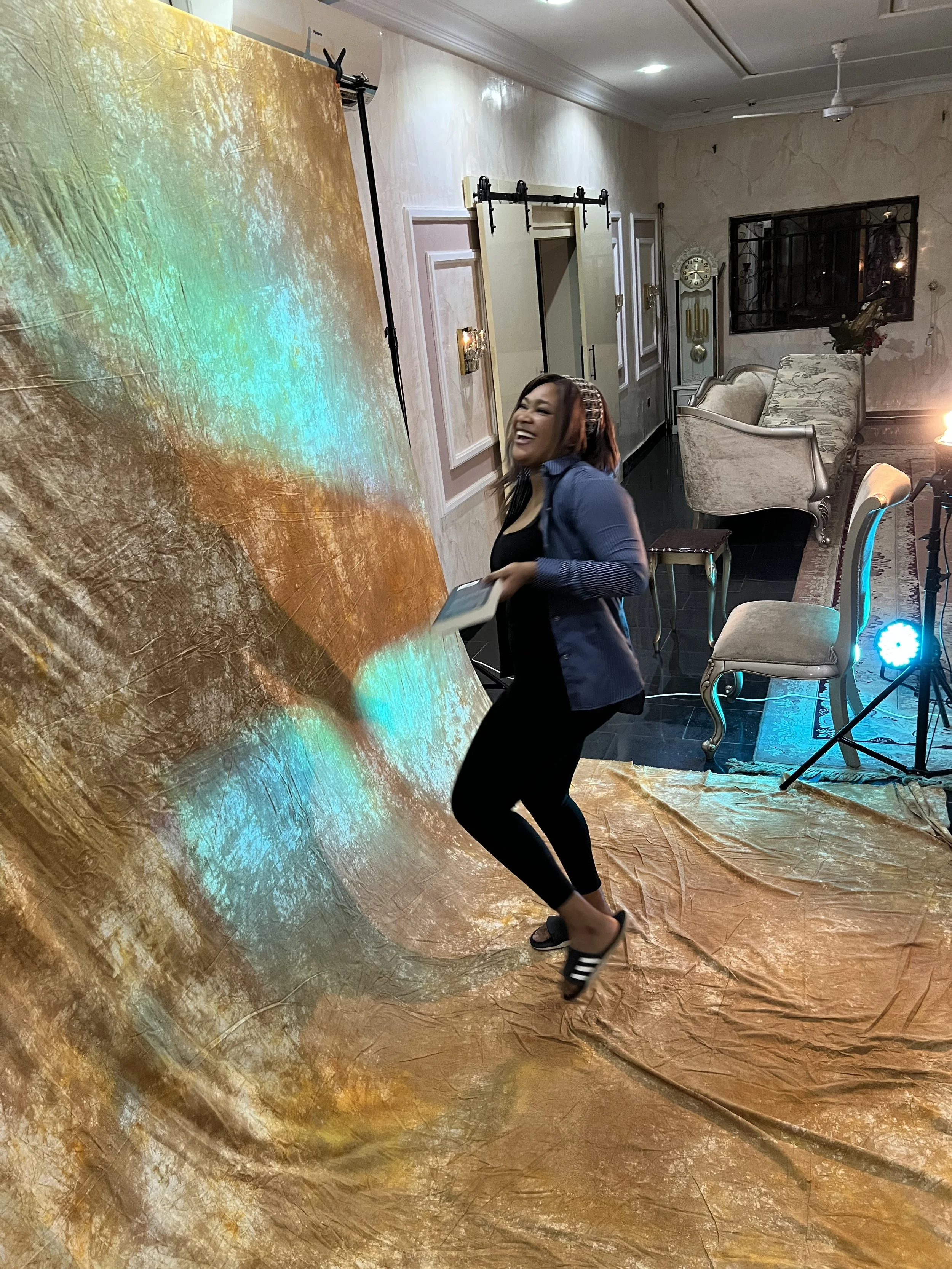 A woman with dark hair, wearing a blue jacket, black top, black pants, and slide sandals, is jumping with joy in front of a large colorful abstract backdrop set up indoors, with furniture and lighting in the background.