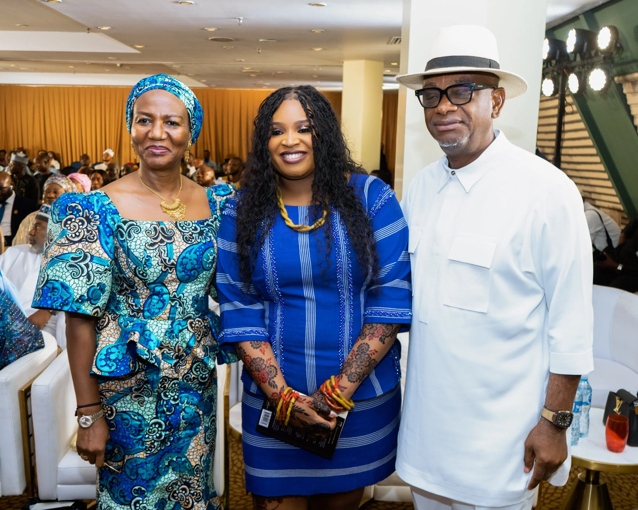 Three adults, two women and one man, are standing together indoors at a formal event, smiling at the camera. The woman on the left is wearing a colorful blue patterned dress with a matching headwrap, the woman in the middle is in a blue dress with traditional Nigerian bead jewelry, and the man on the right is dressed in a white traditional suit with a white hat and black glasses.