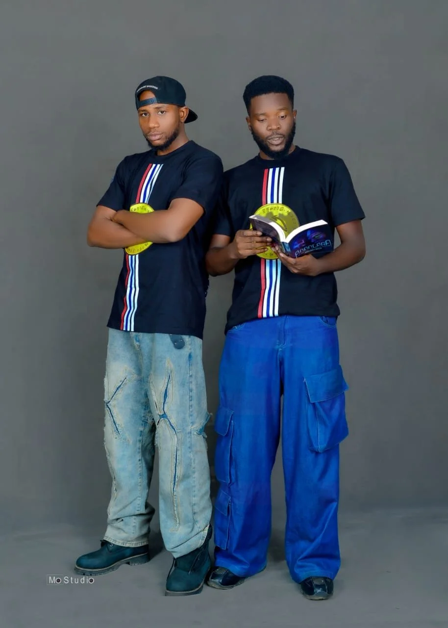 Two young men stand against a gray backdrop, wearing matching black T-shirts with red, white, and blue vertical stripes and yellow circular logos. One man on the left has a cap backward, arms crossed, and is wearing distressed jeans and boots. The man on the right is holding an open booklet or magazine, has a beard, and is wearing blue cargo pants and black shoes.